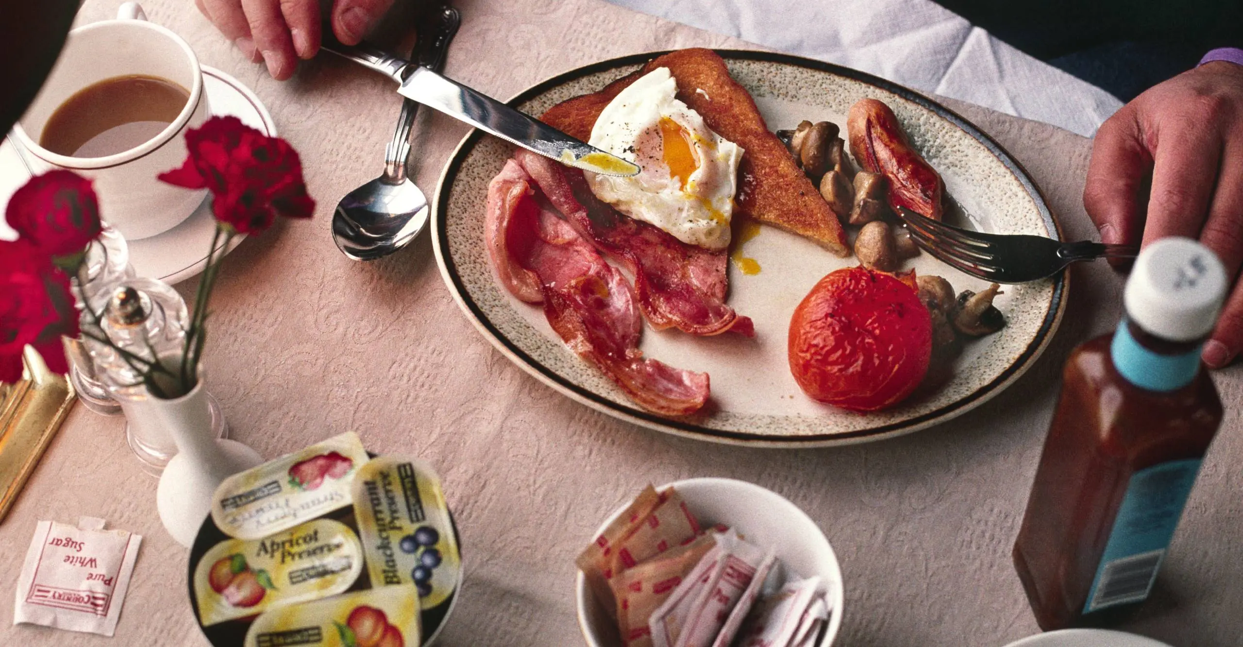 Colour photograph of a table set with someone eating a cooked breakfast of bacon, egg, sausage and tomato