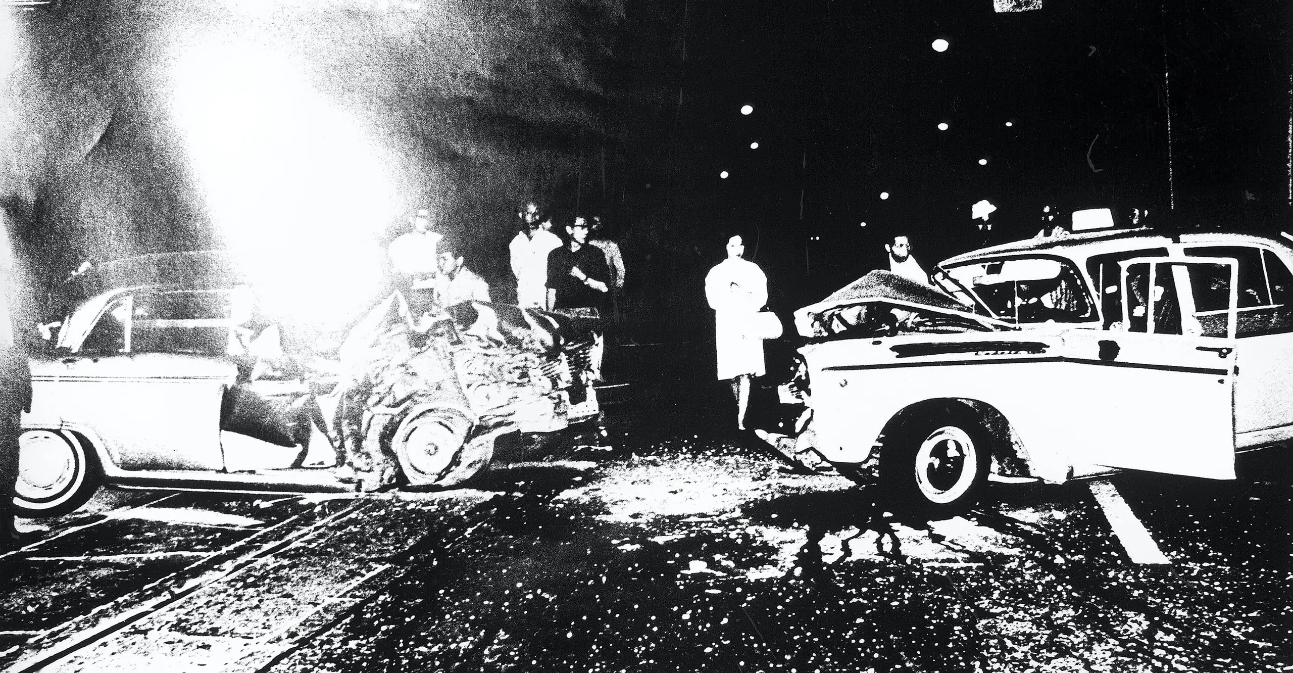 High contrast black and white photograph of two severely damaged cars and a small crowd