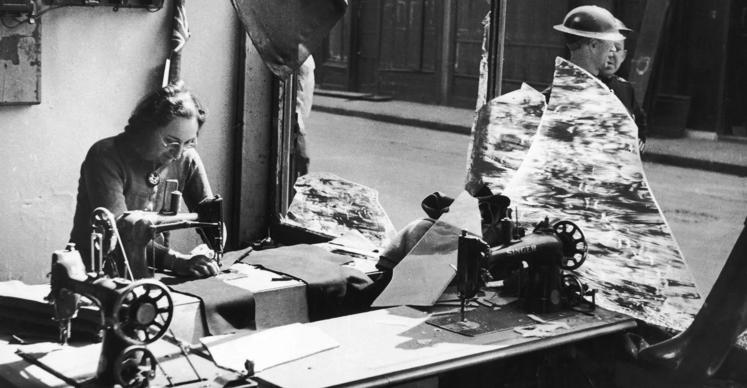 Black and white photograph of a woman working amongst the broken glass of a tailor’s shop