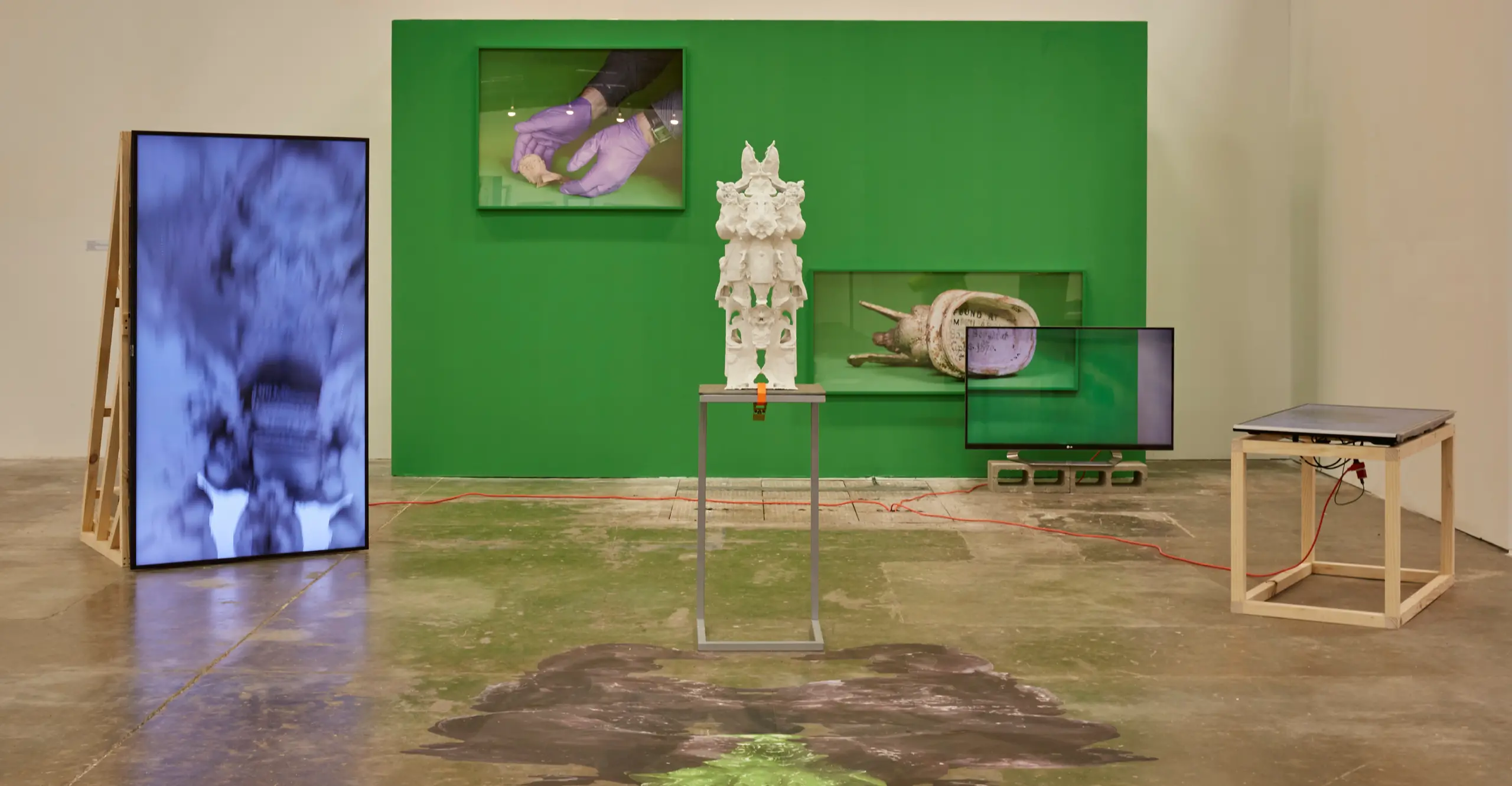 An installation view of an exhibition on a gallery. There's a sculpture in the middle of the image, a built wall painted in green in the back, a large vertical screen on the left and other art works on view.