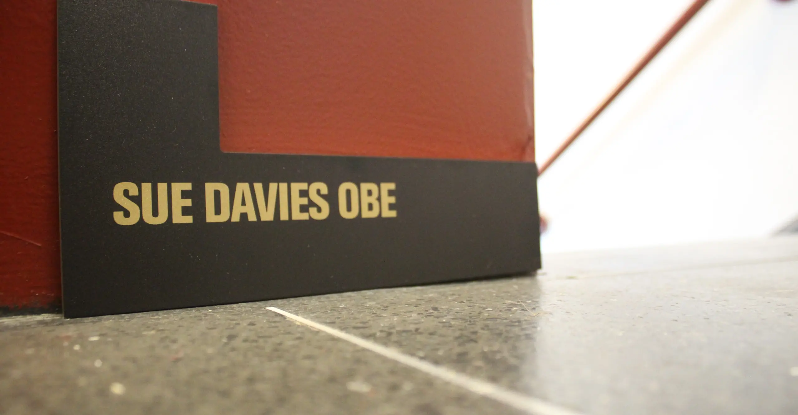A black L shape with gold writing saying 'Sue Davies OBE'