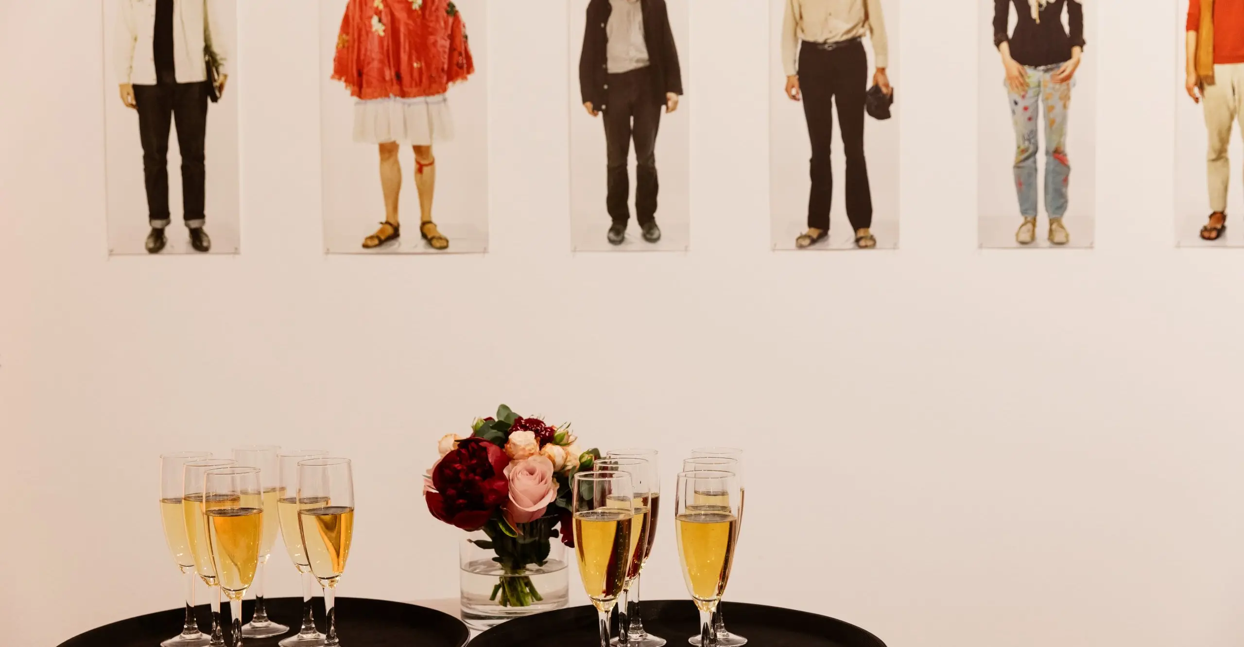 A photograph of two trays with champagne glasses on them, a bunch of flowers and an exhibition in the background