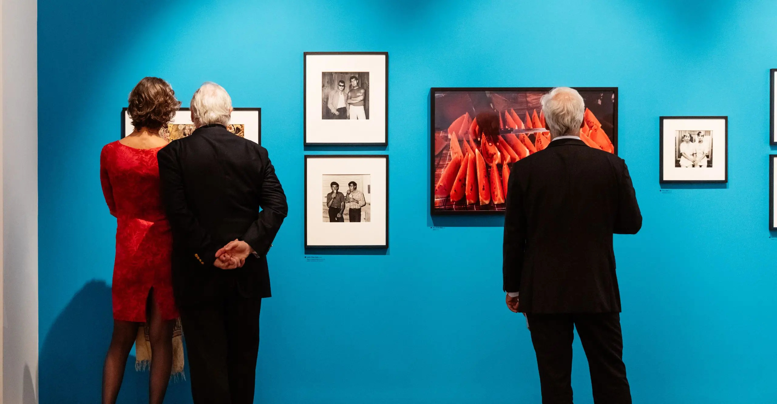 Colour photograph of three people with their backs to the camera, looking at a blue wall with framed photographs