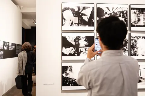 Colour photograph of a person standing with their back to the camera, holding up a phone taking a picture of black and white photographs on the wall