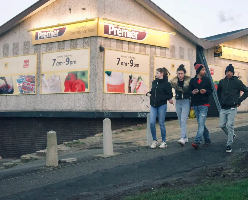 Four young people walk in a row down a paved hill past a Premier convenience store at dusk.