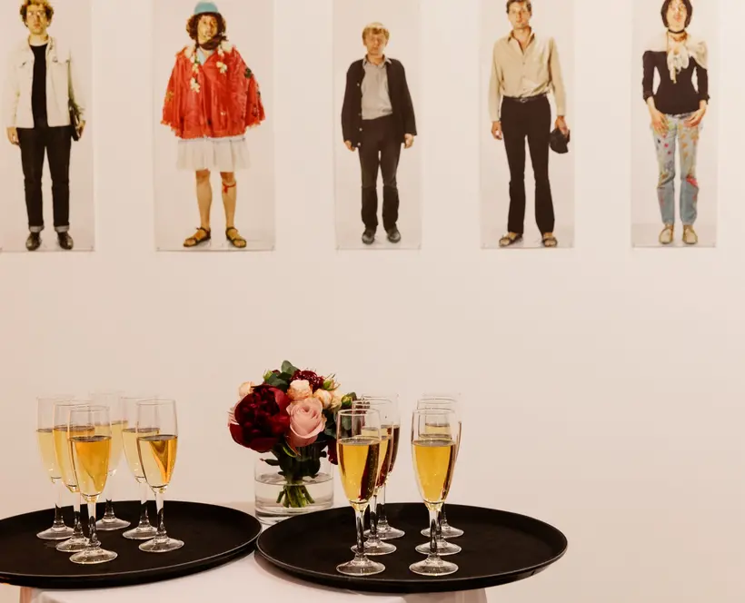 A photograph of two trays with champagne glasses on them, a bunch of flowers and an exhibition in the background