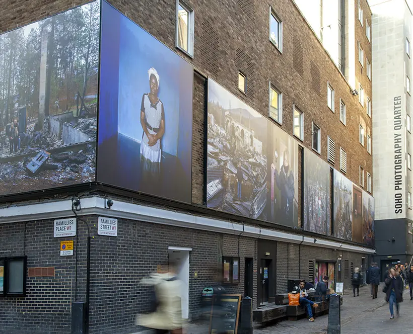 A frieze of colour photographs on an outdoor building depicting portraits of people standing in the aftermath of a fire or flood.
