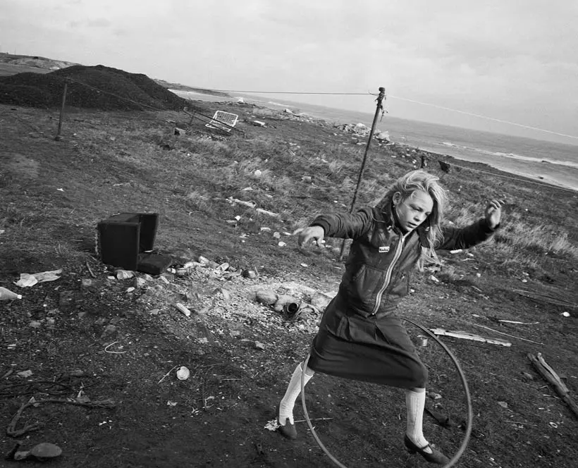 Black and white photograph of a girl playing with a hula-hoop, against a deindustrialized landscape