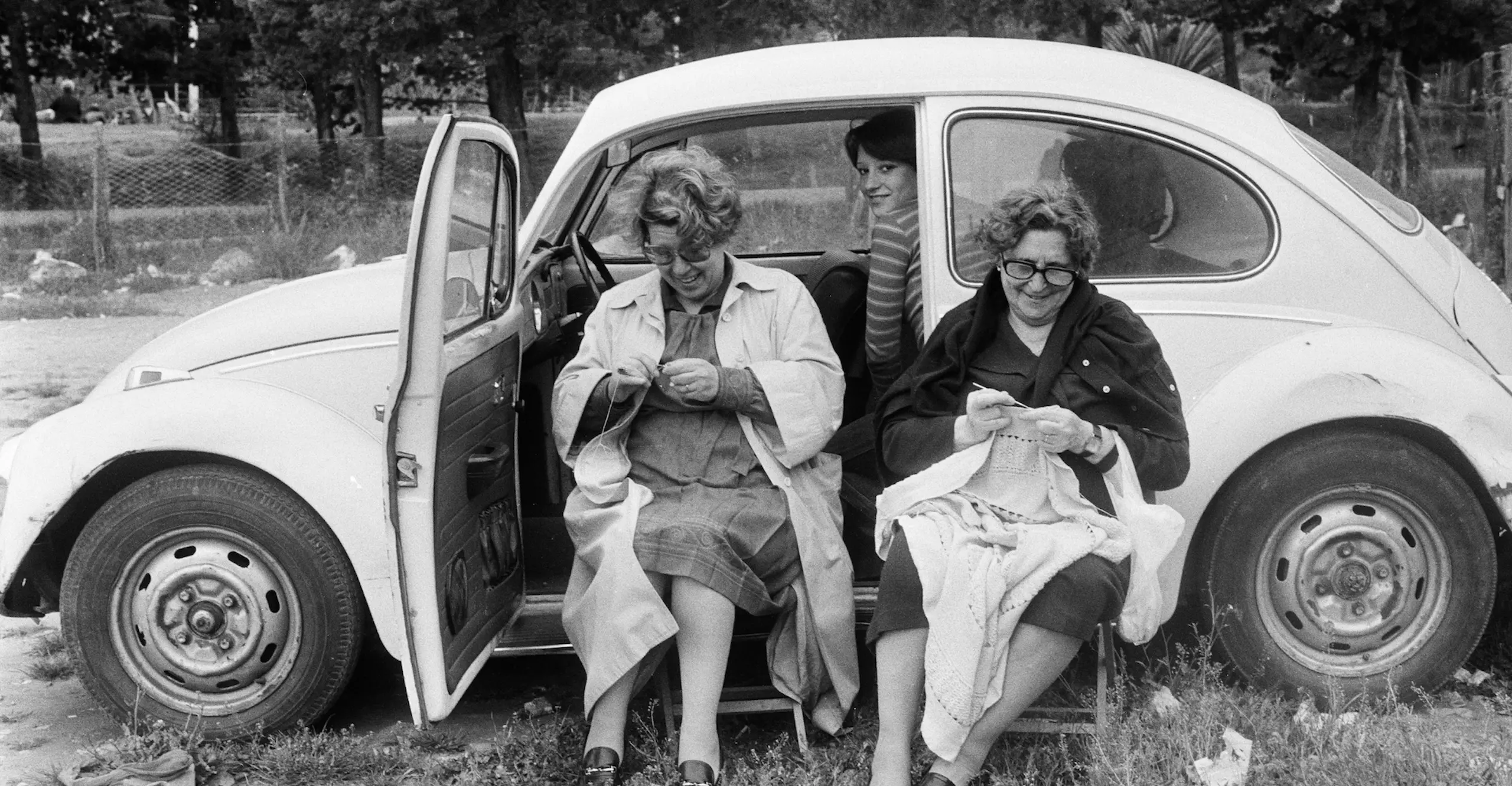 Black and white photograph of two women sat outside a car. They are both busy crocheting.