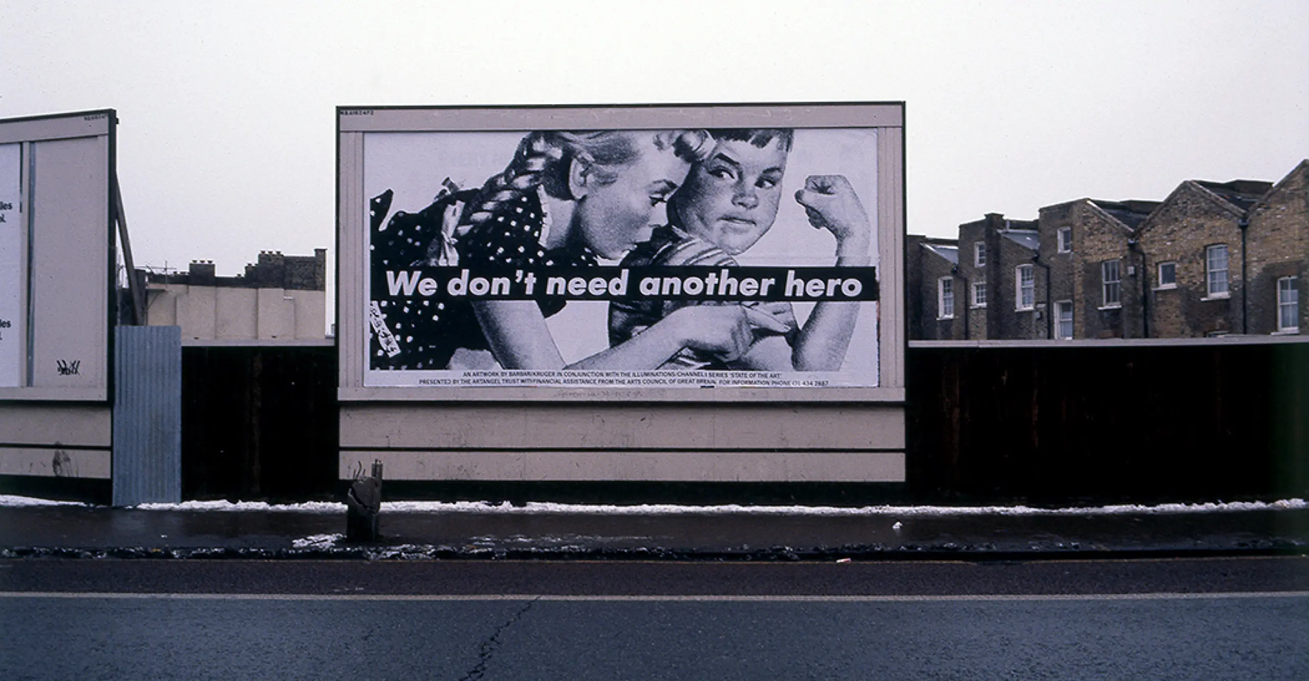Black and white photograph of a billboard featuring a vintage-style advert of two children and the words "we don't need another hero" printed across