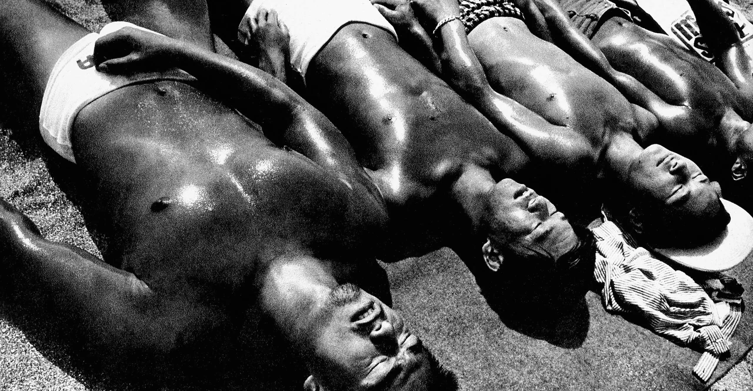 Black and white photograph of a group of men sunbathing, laying in a row next to each other wearing swimming trunks