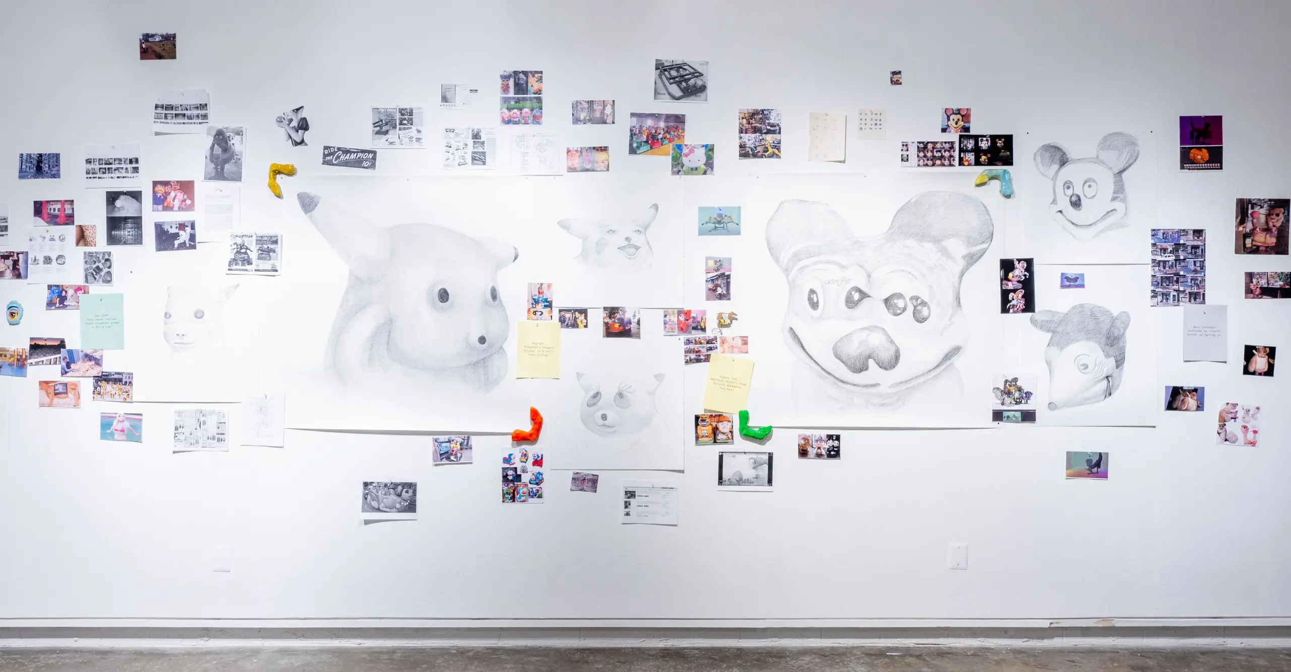 A photograph of white wall with several small photographs glued and a few hand painted creatures painted with pencil