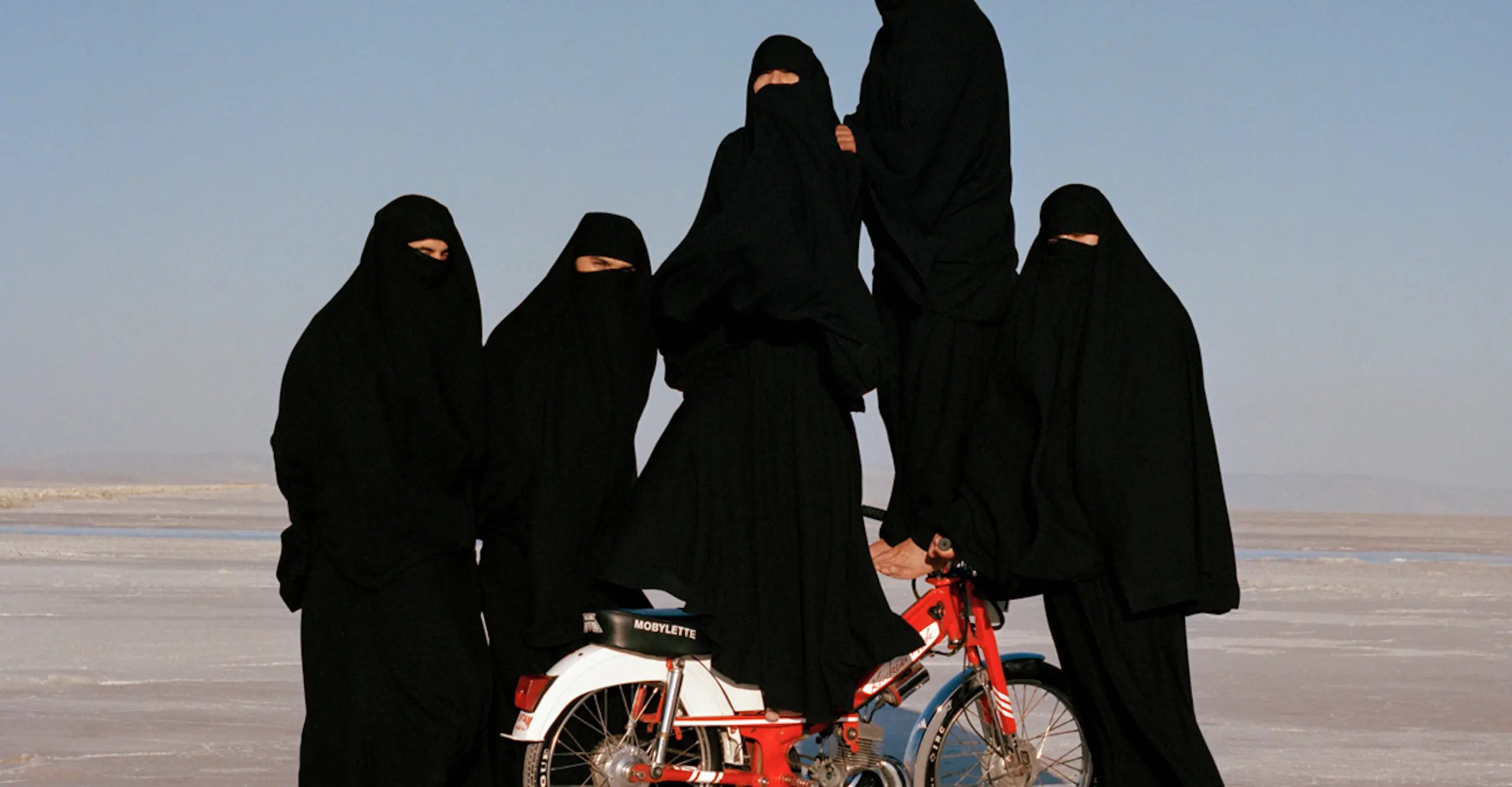 Five figures dressed in black hijabs stand in the desert with two balanced on a motor scooter.