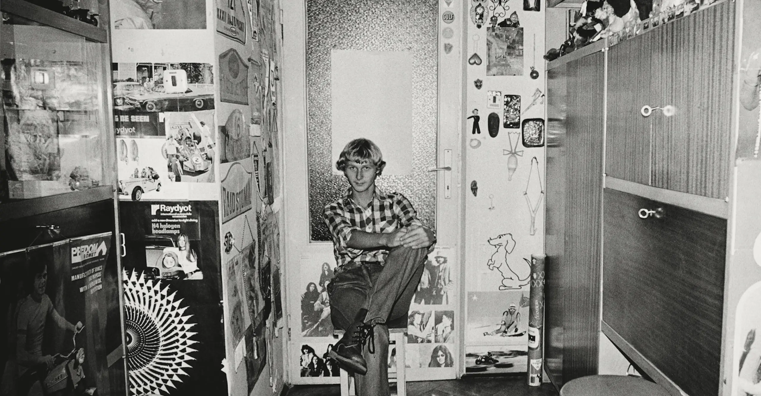 Black and white photograph of a young man  sat on a stool in a room full of signs and stickers