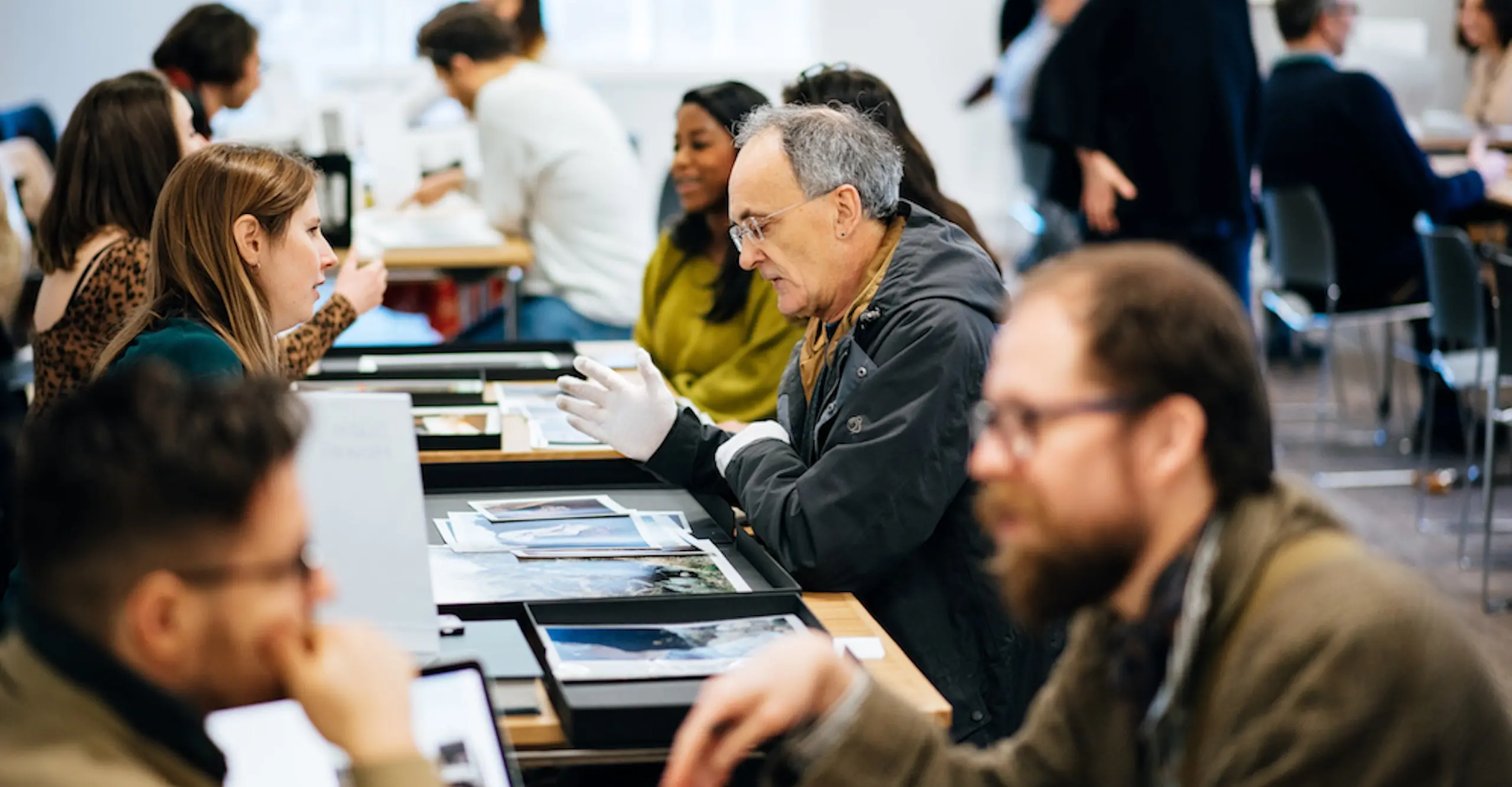 Photograph of people sitting across from each other at tables talking. Portfolios of photographs are on the tables.