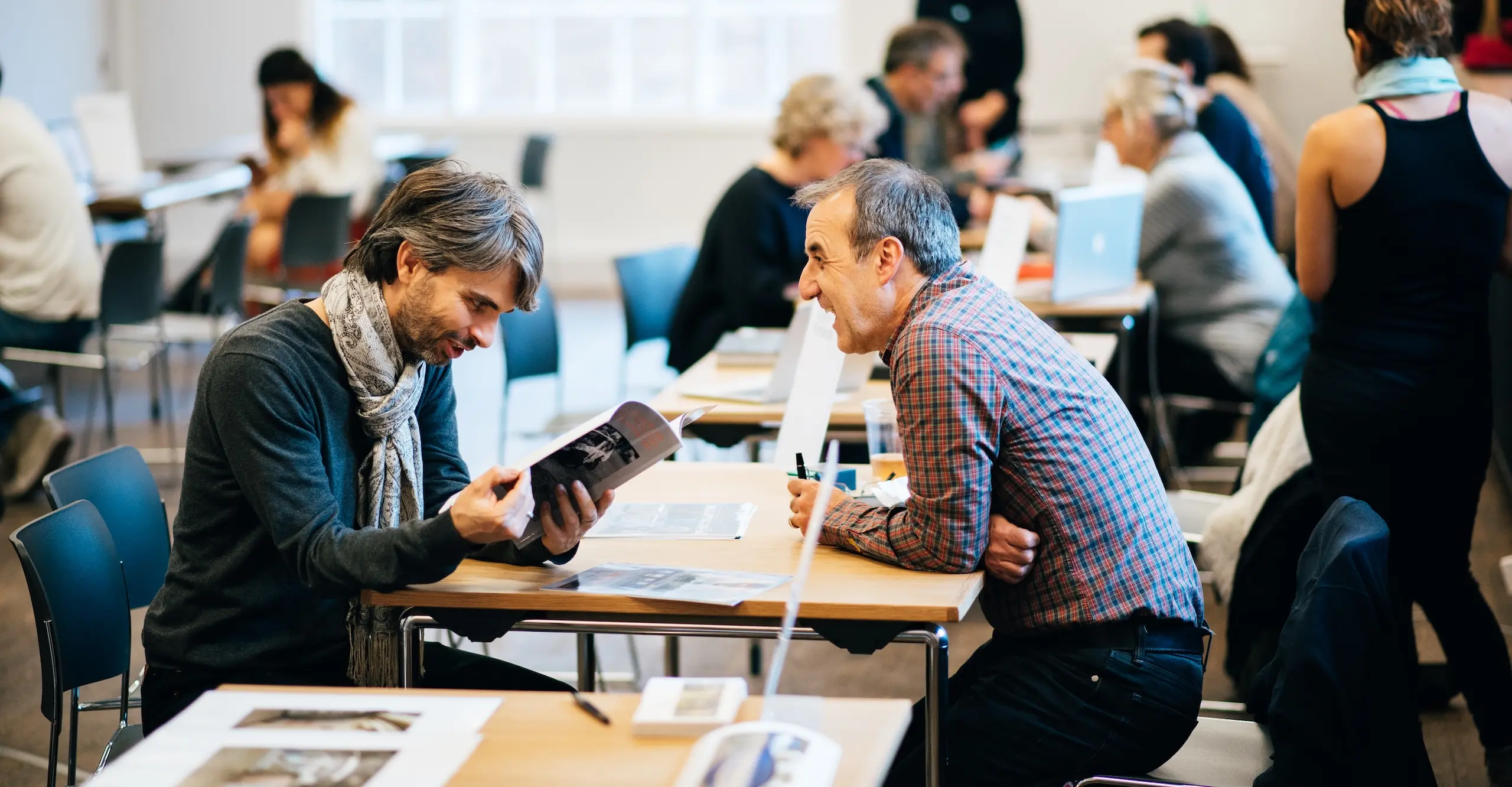 People sitting across from each other and talking at tables with photographs or laptops on the tables.