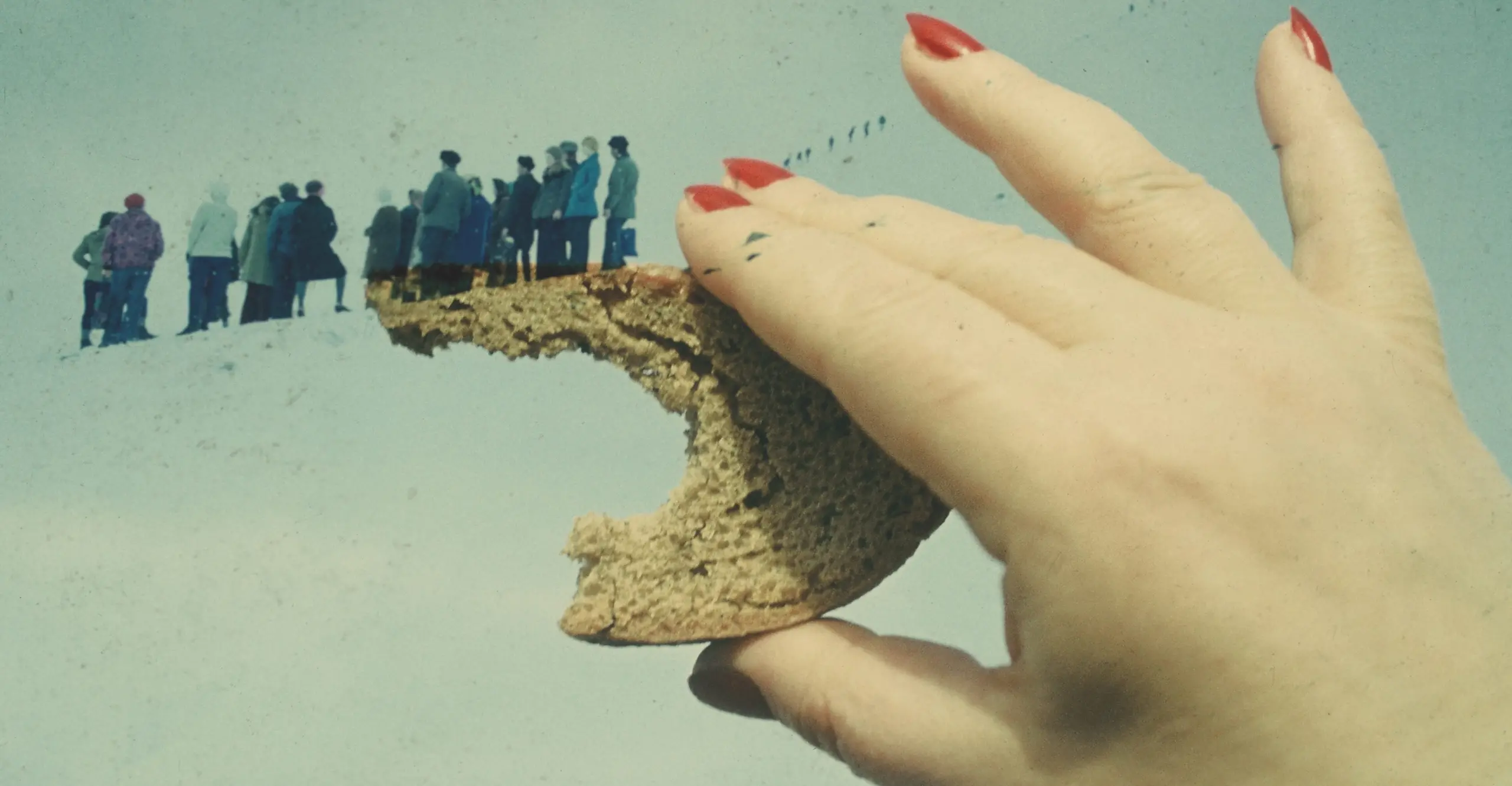 Colour photograph of a close-up hand  holding a half-eaten piece of bread. In the back of the image, a group of people in winter coats can be seen huddled together