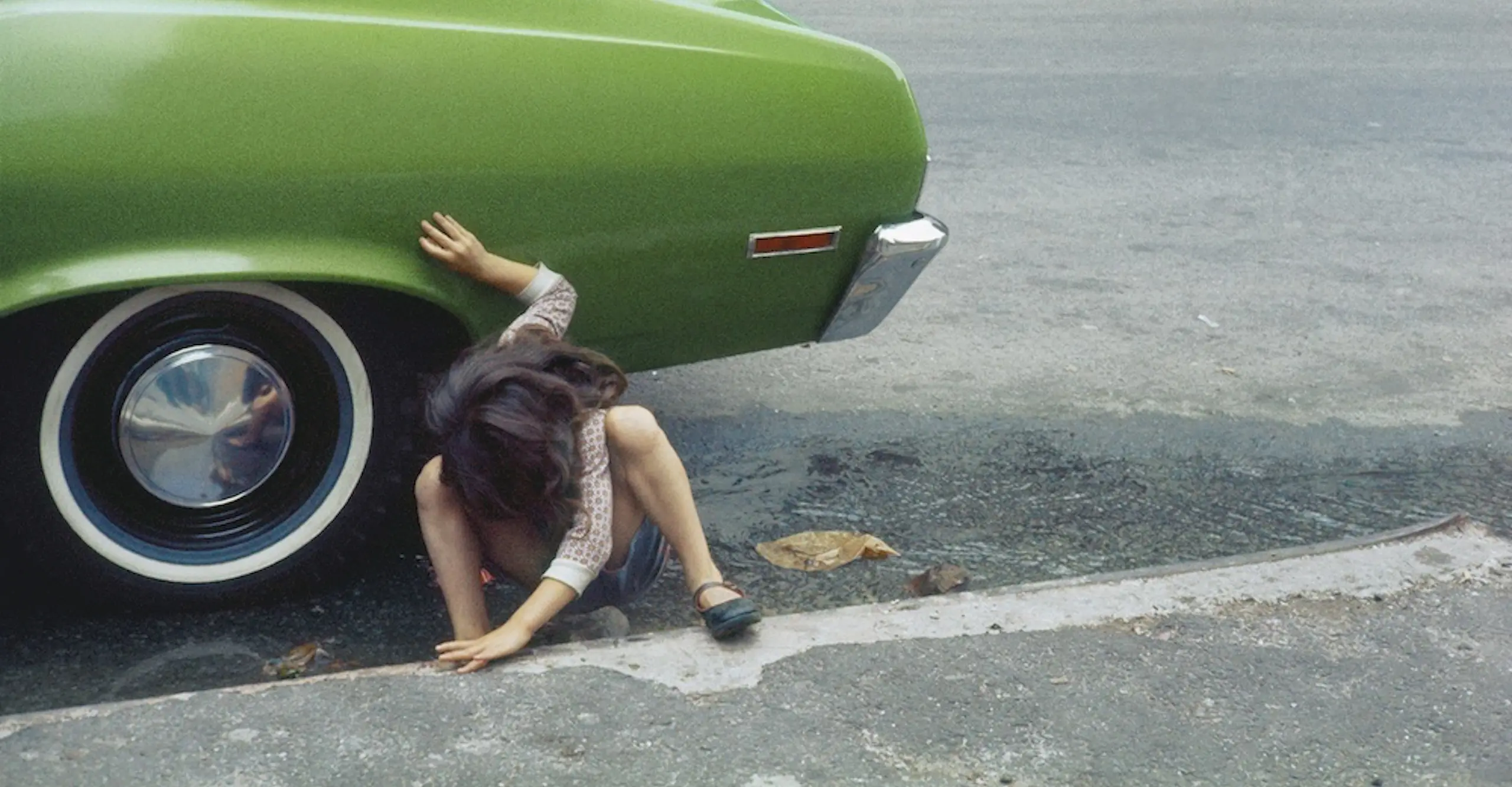 A girl squats beside the rear tyre of an avocado green car, body and face twisted away from the camera.