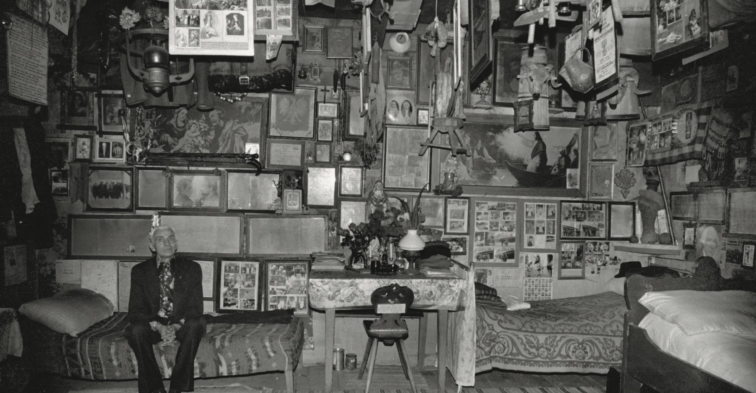 Black and white photograph of an older  man sat on a bed in a bedroom filled with numerous framed  pictures, religious images and objects hanging from the  walls and ceiling.