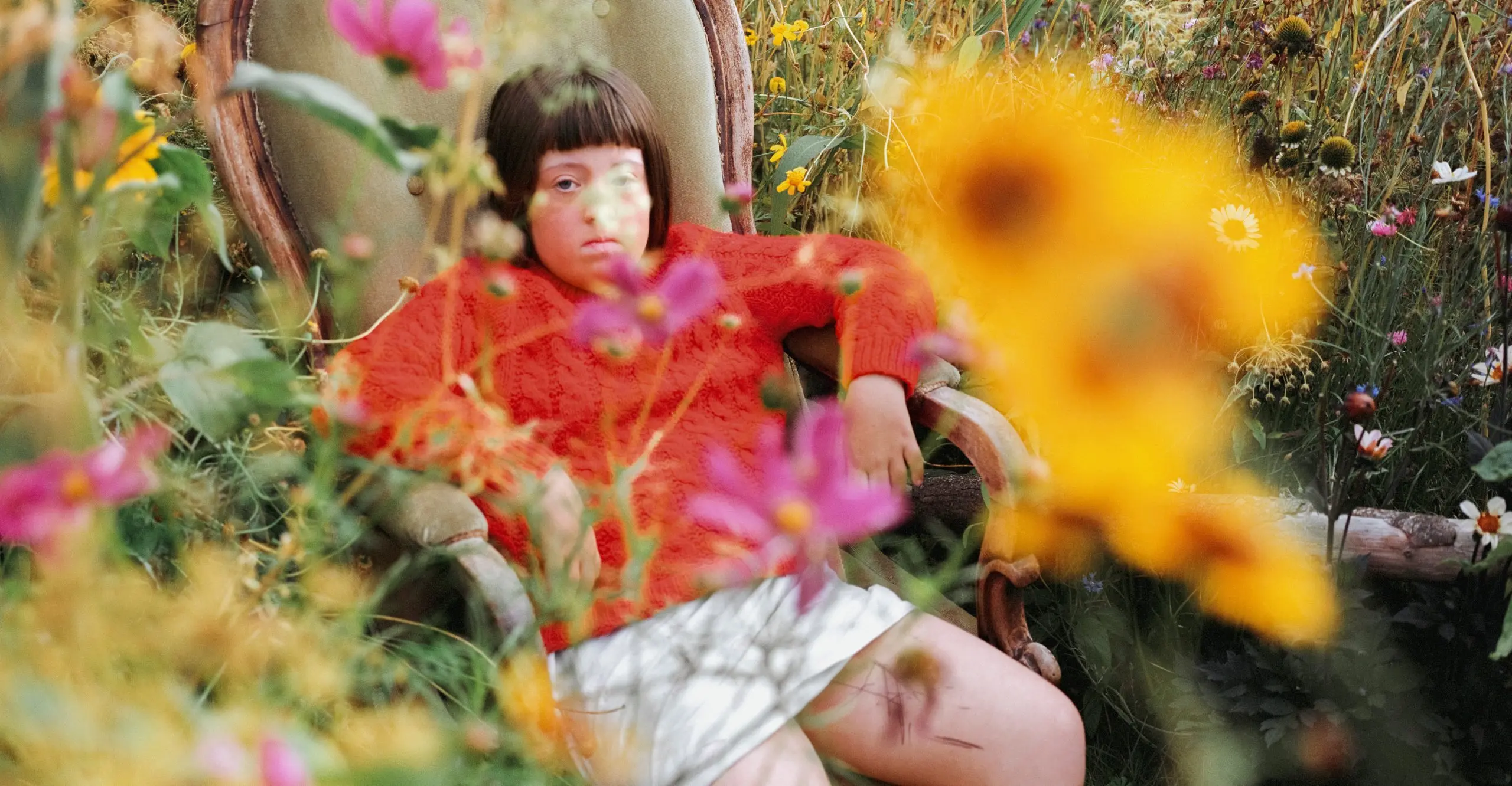Colour photograph of a young person sat in an armchair in the middle of a garden with flowers in the foreground
