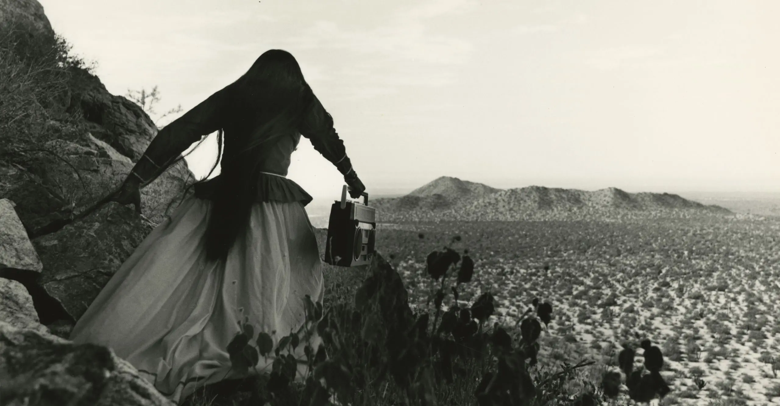 Black and white image of a woman walking in the desert, her back is to the camera as she walks away