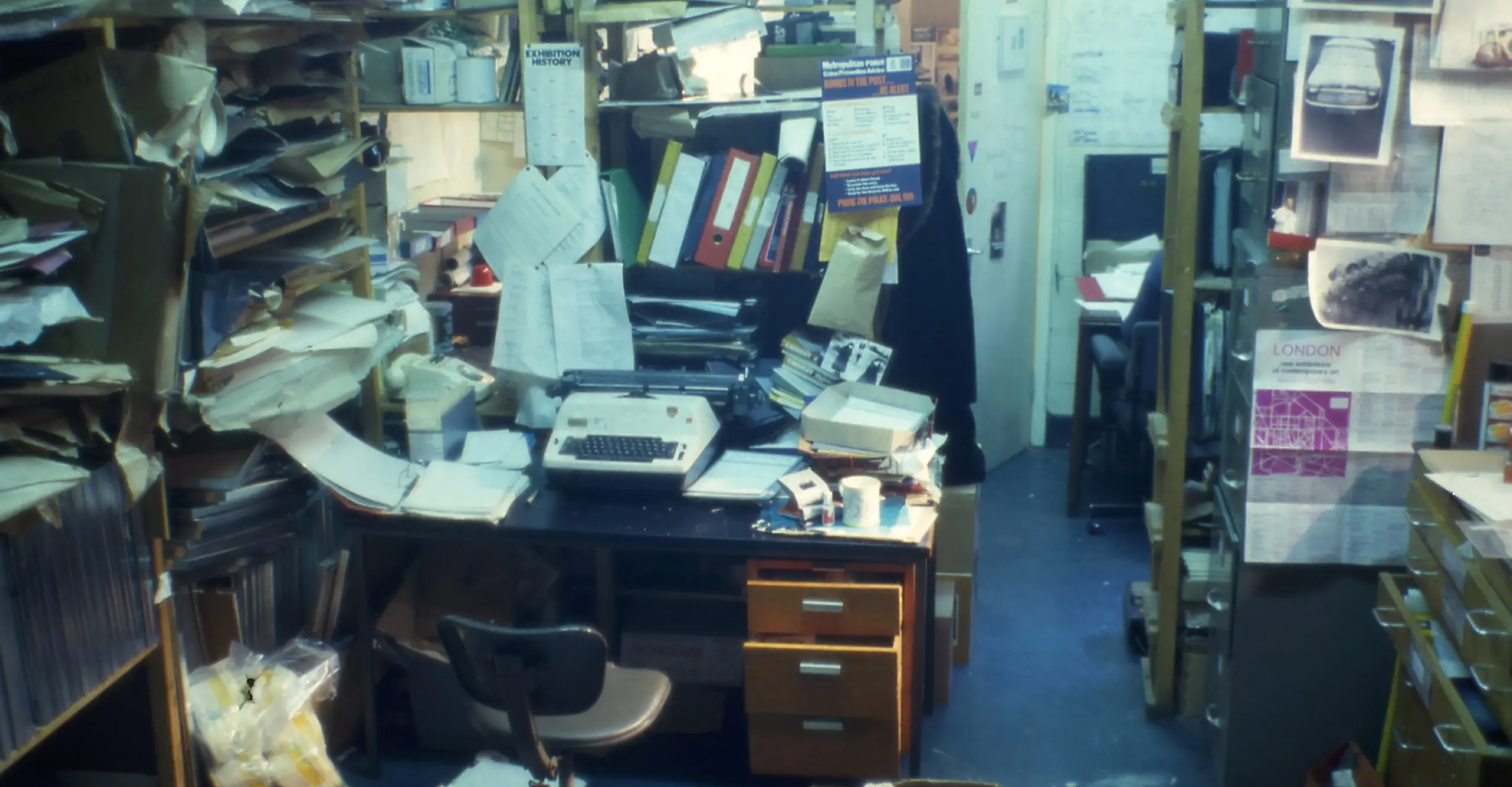 Archival full colour photograph with a desk in the middle of an office surrounded by papers, books, binders and a miscellany of other office supplies and materials
