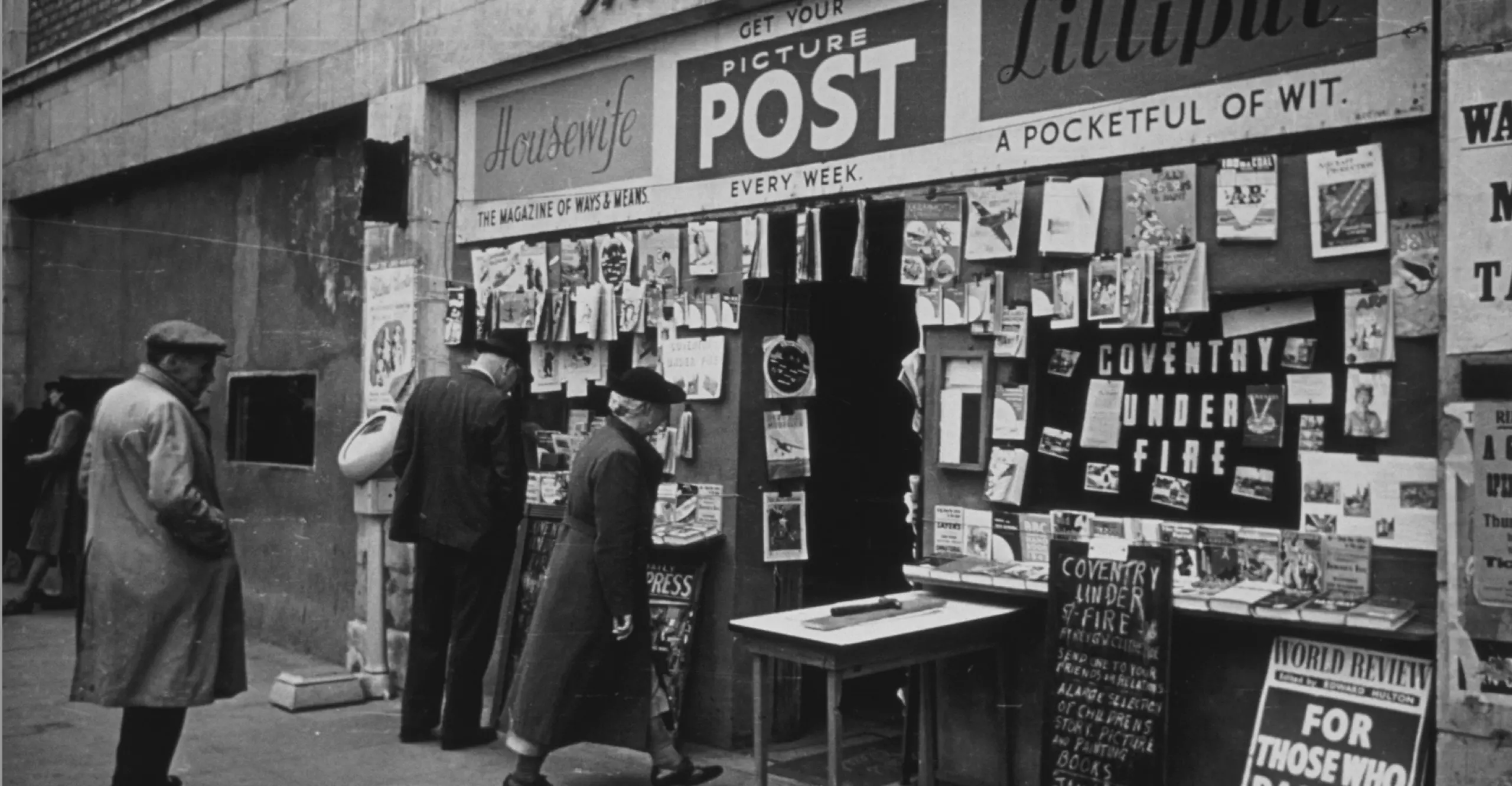 Black and white image showing a news stand with the words "Picture Post" and several figures standing outside