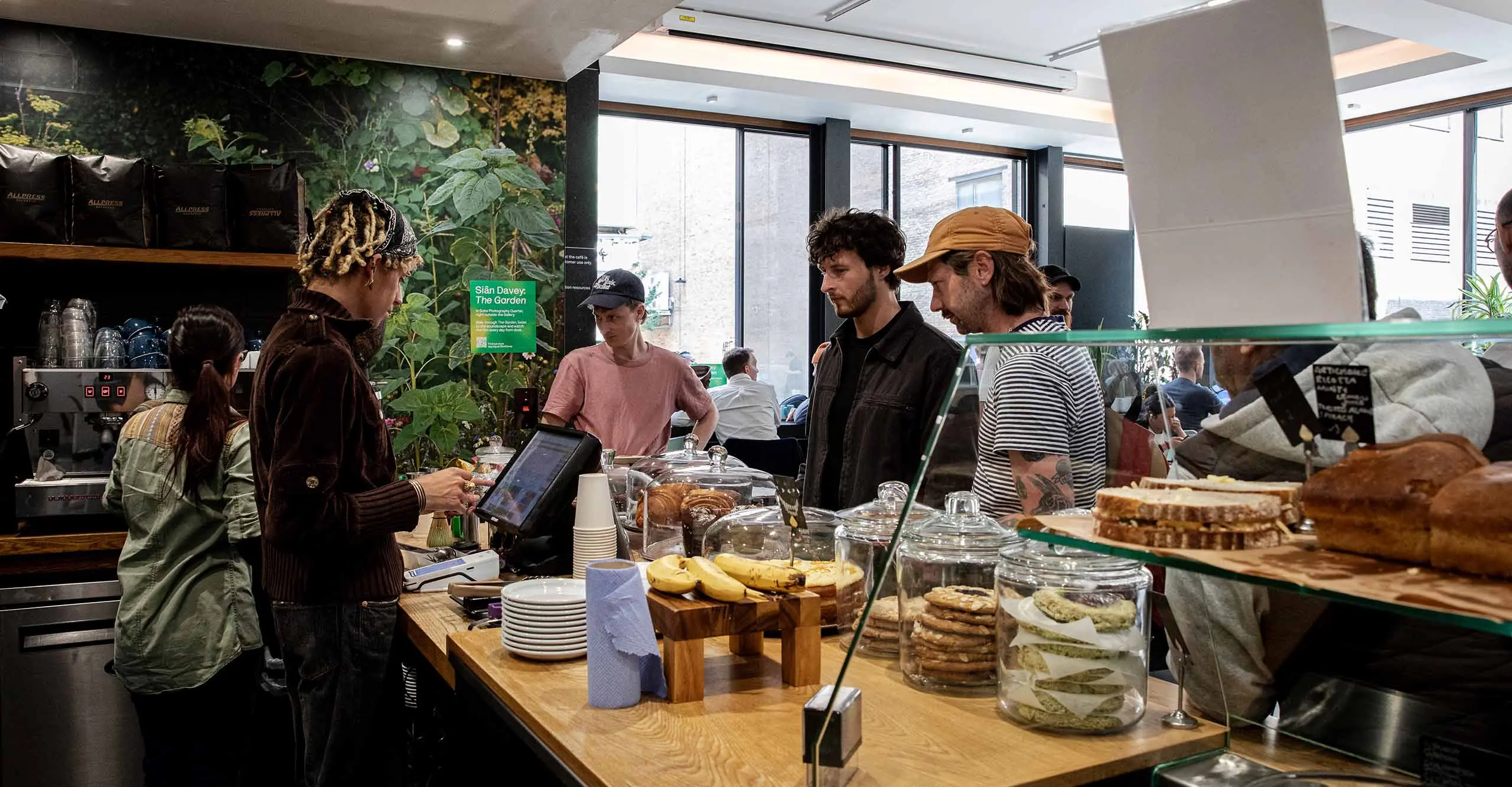 Image of The Photographers' Gallery Cafe. Customer is in the left of the image, with their face turned away, wearing a light denim shirt, being given a coffee cup and saucer on the counter by the man stood next to the coffee machine in the cafe.