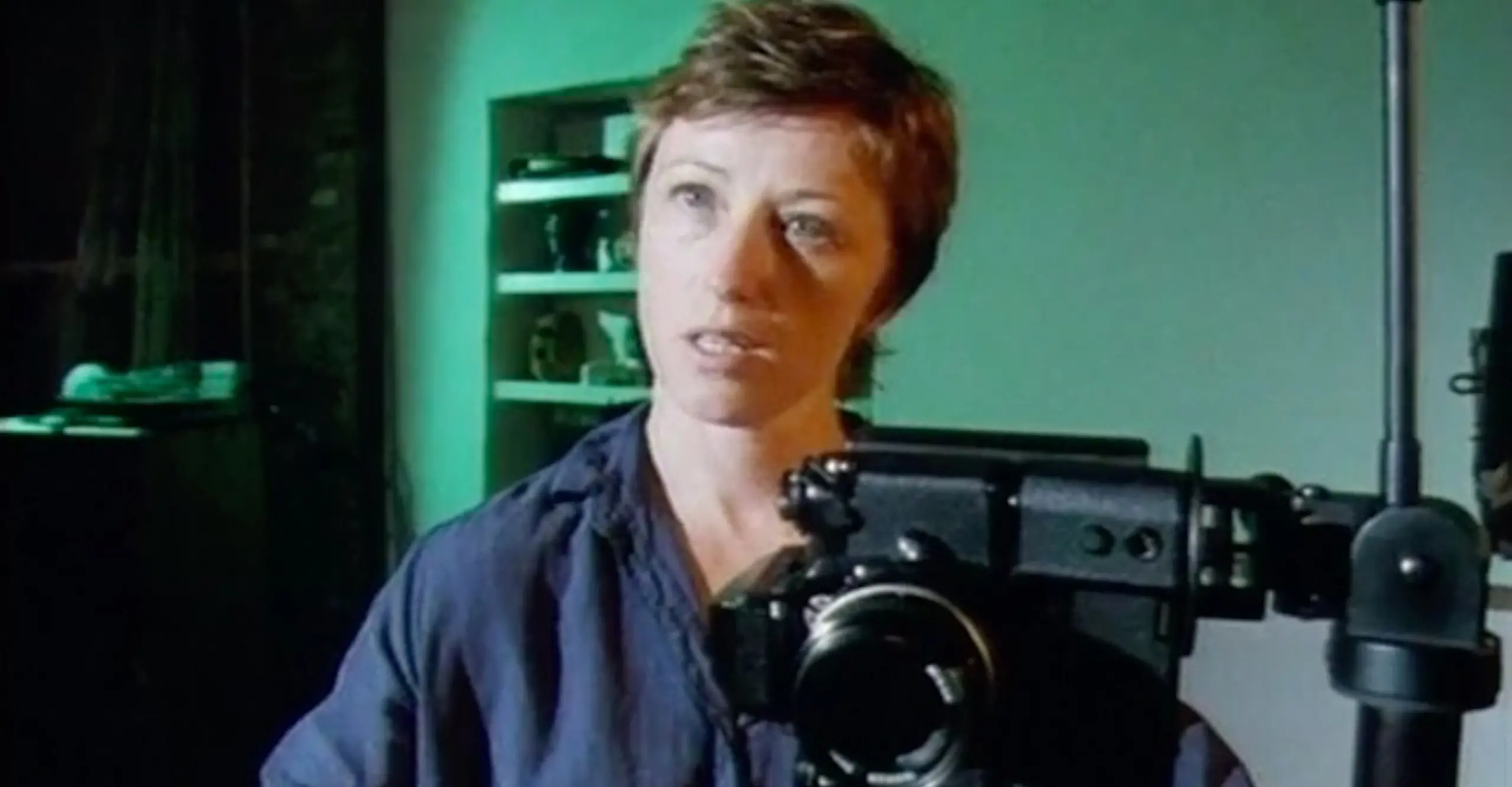 Head and shoulders of a white woman with cropped hair and a camera in the foreground and a green wall behind.