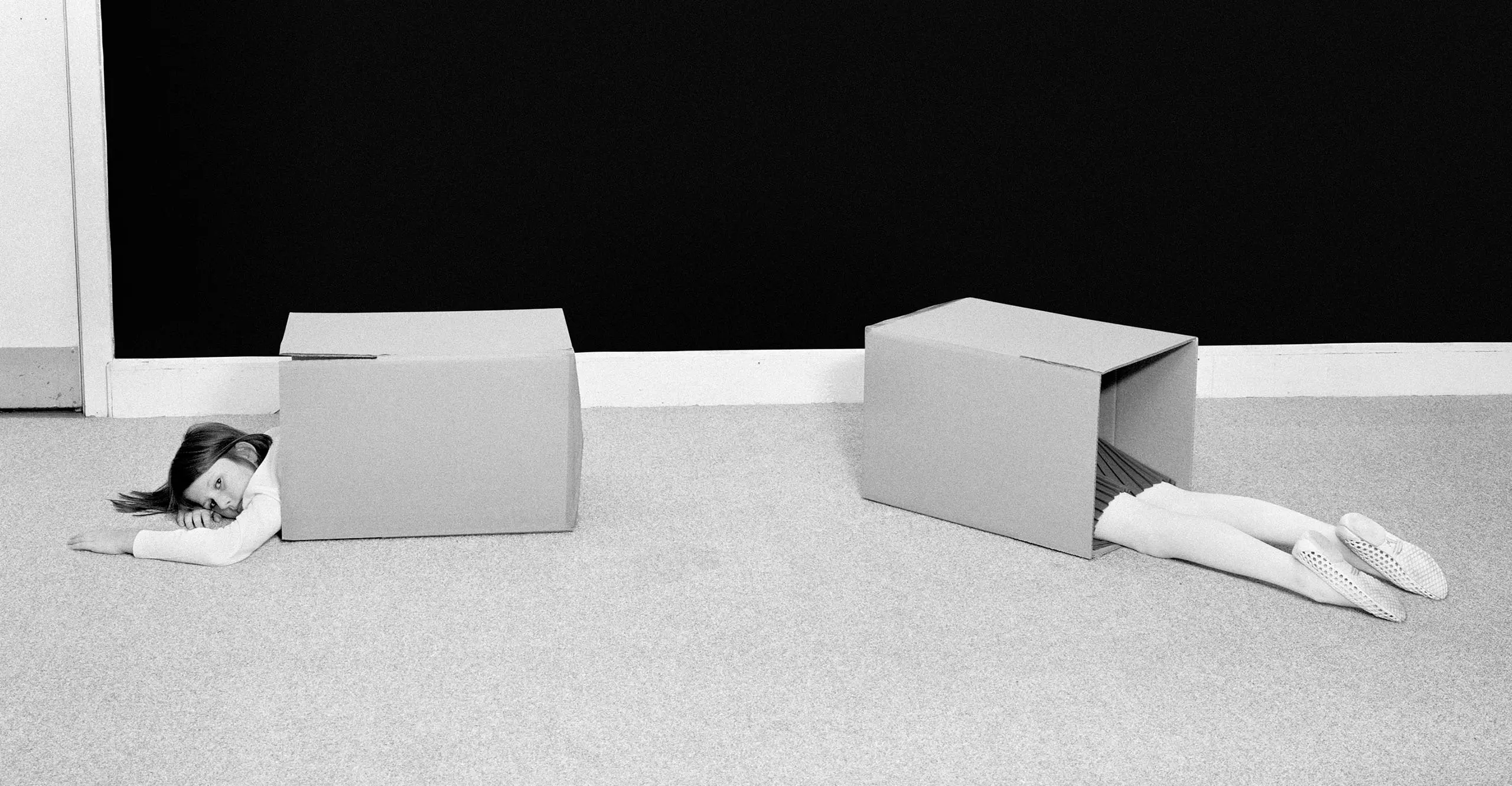 Black and white image of a girl lying on the floor with 2 boxes. Half of her torso is showing in one box and the lower half of her is showing in another box