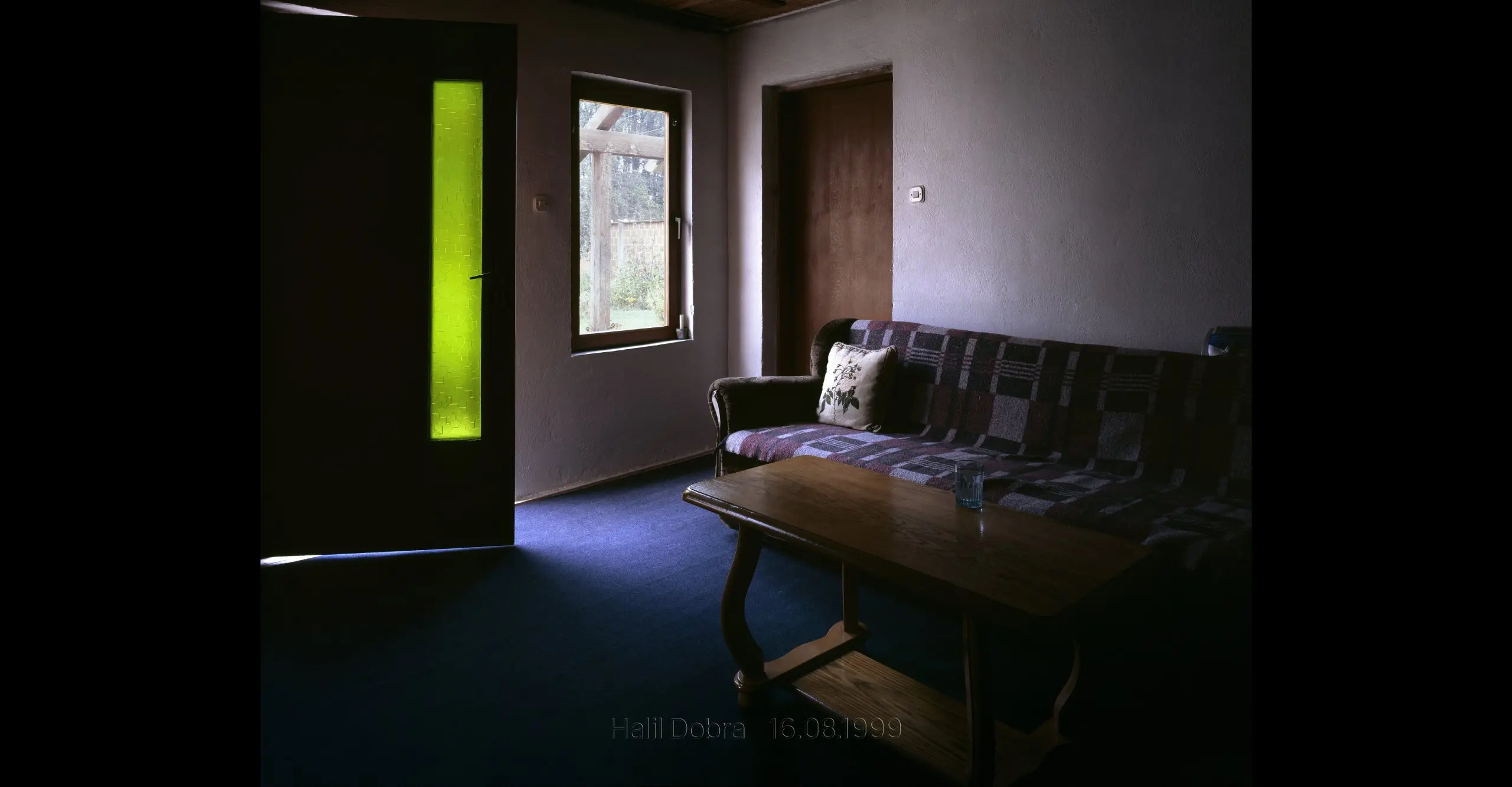 Colour photograph of a darkened room  with a sofa, table and a door with green glass, the image  states ‘Halil Dobra 16.08.1999’