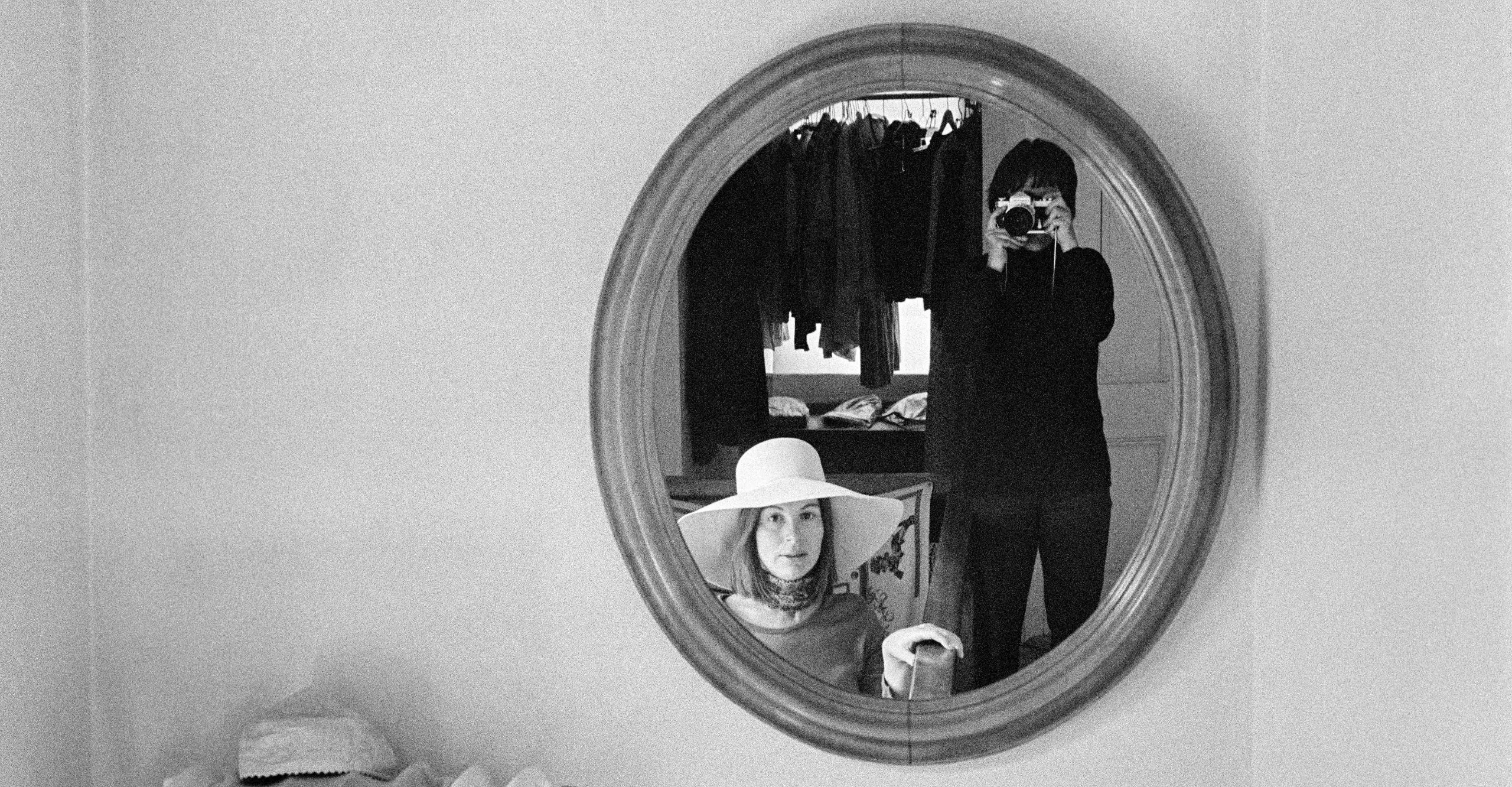 Black and white photograph showing a mirror on a plain wall. Reflected in the mirror is a woman wearing a wide brimmed hat and stood next to her, a woman holding up a camera to take the photo, wearing a black top and trousers