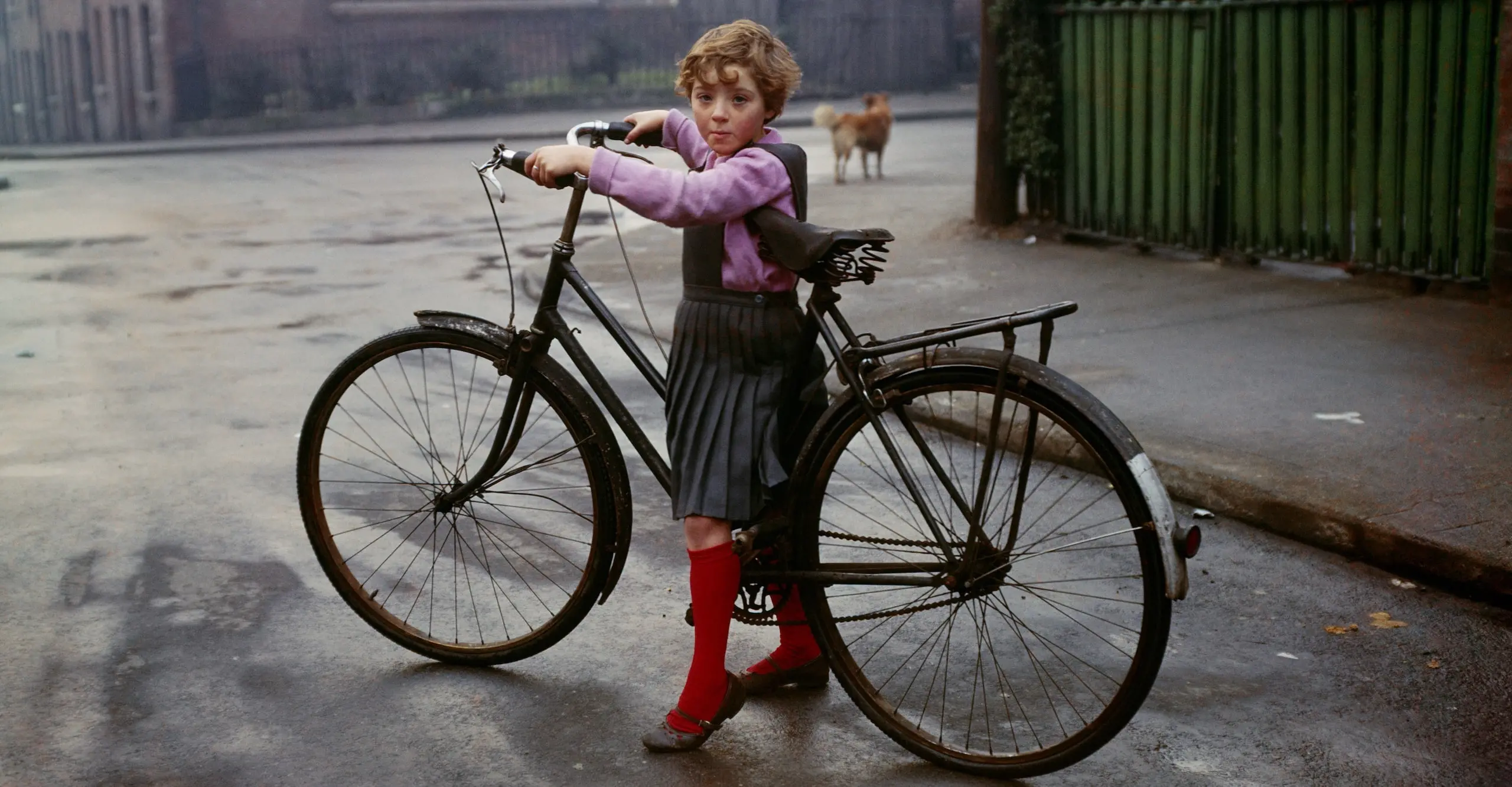 Colour photograph of a small child, straddling the frame of an adult-sized bicycle, who has stopped on a street and turned to look at the photographer.