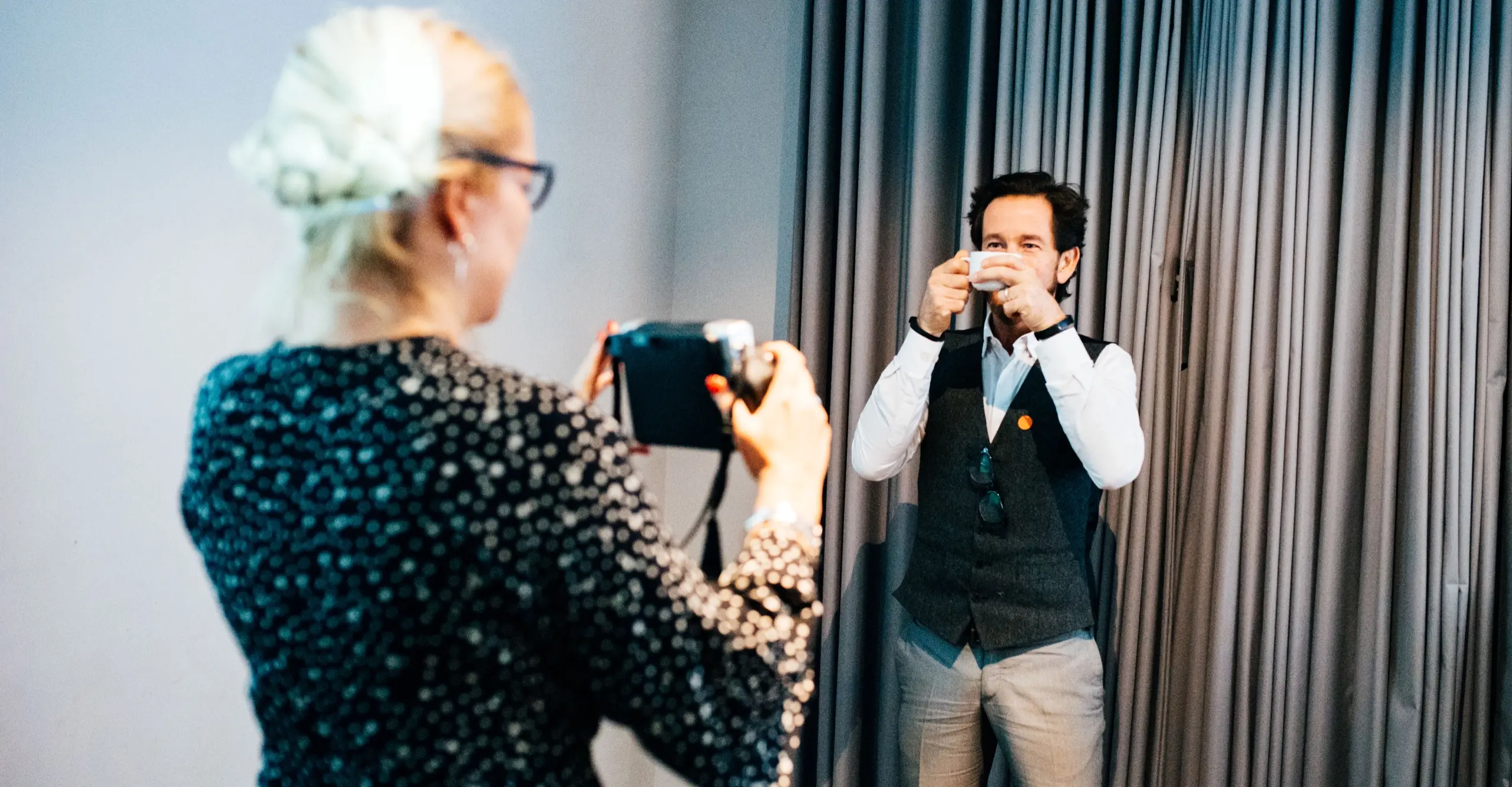 Photograph of white woman taking a photo of a man holding a cup in front of his face while standing in front of a grey curtain