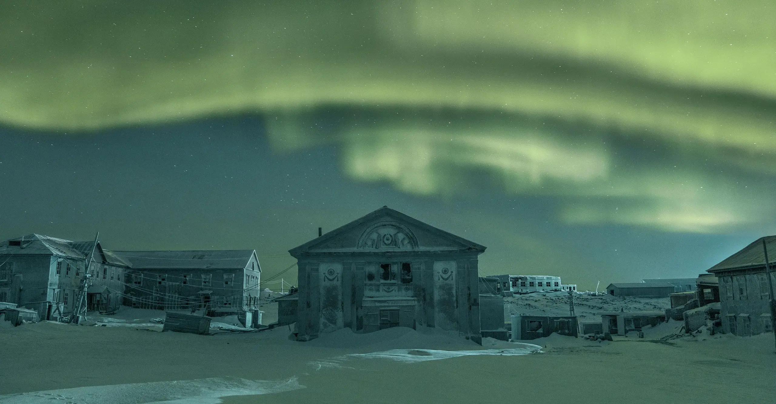 Colour photo of a snowy landscape bathed in a green glow from the overhead northern lights. A house is in the centre.