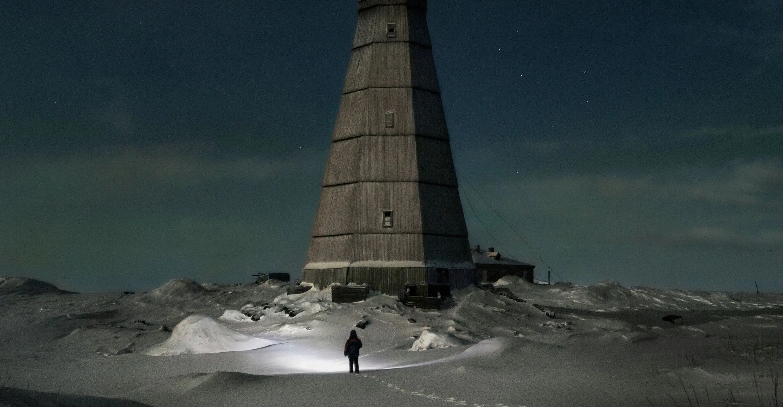 Dark, atmospheric colour photograph of a snow landcape at night with a silhouetted figure standing infront of a large lighthouse building