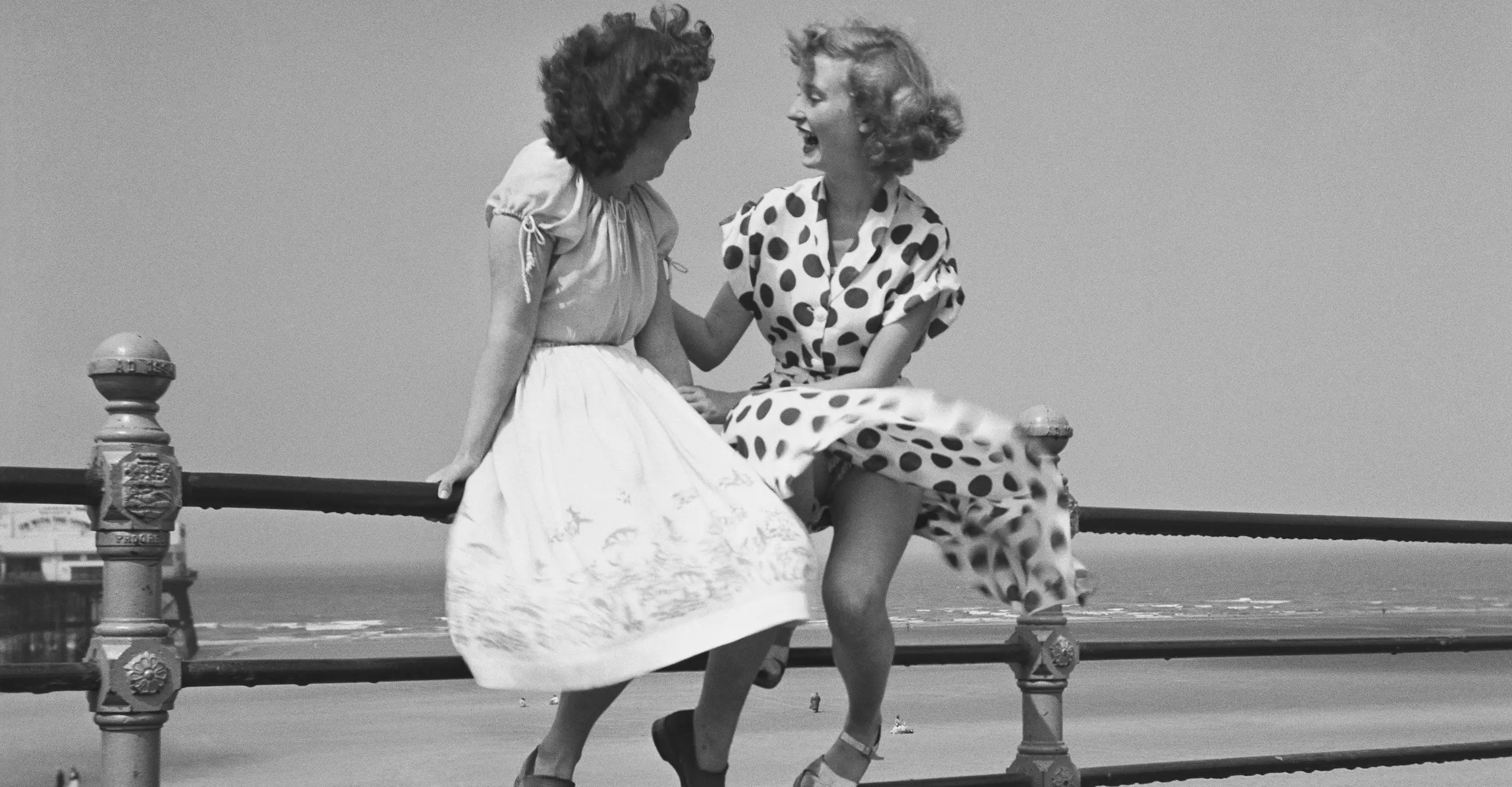 Black and white photo of two womenchatting on the railings by a beach.