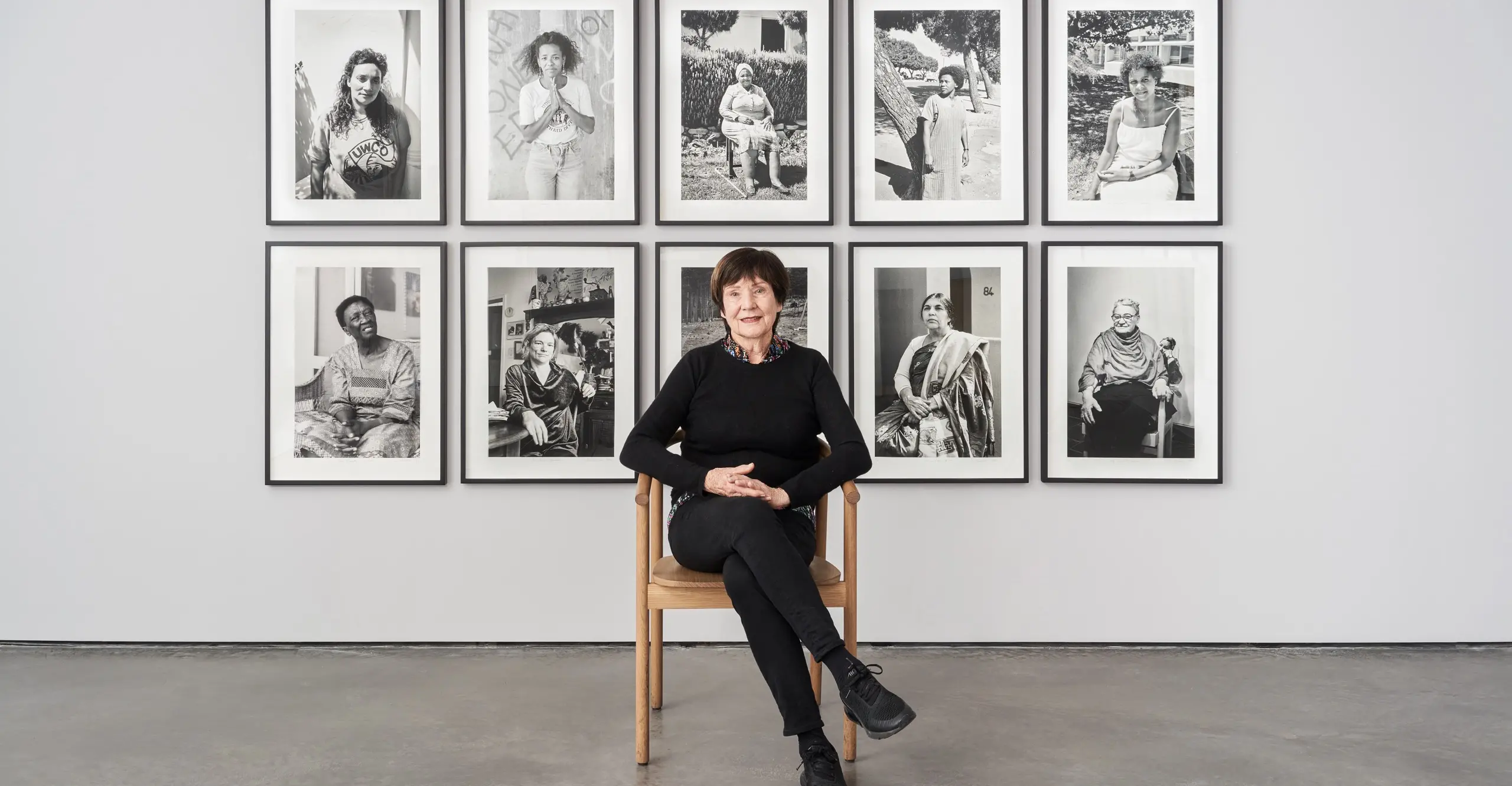 Portrait of the artist wearing all black, seated in front of an installation of 10 B&W images by the artist