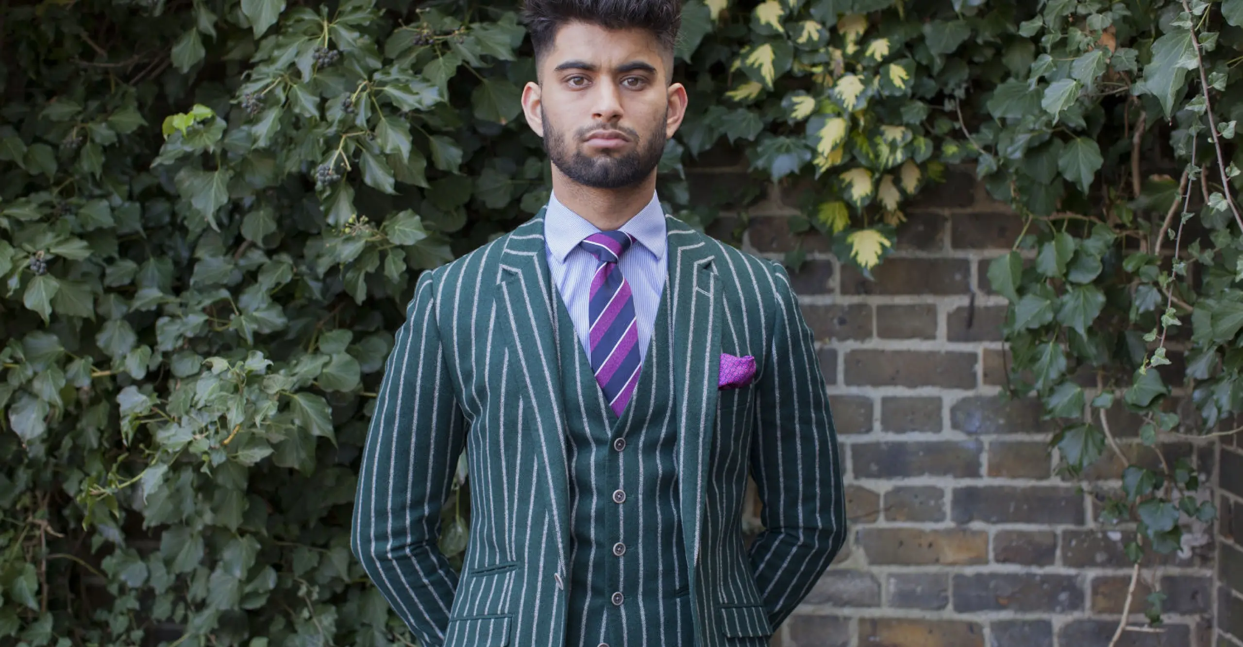 Landscape format portrait of a man wearing a green striped suit with a brick wall in the background