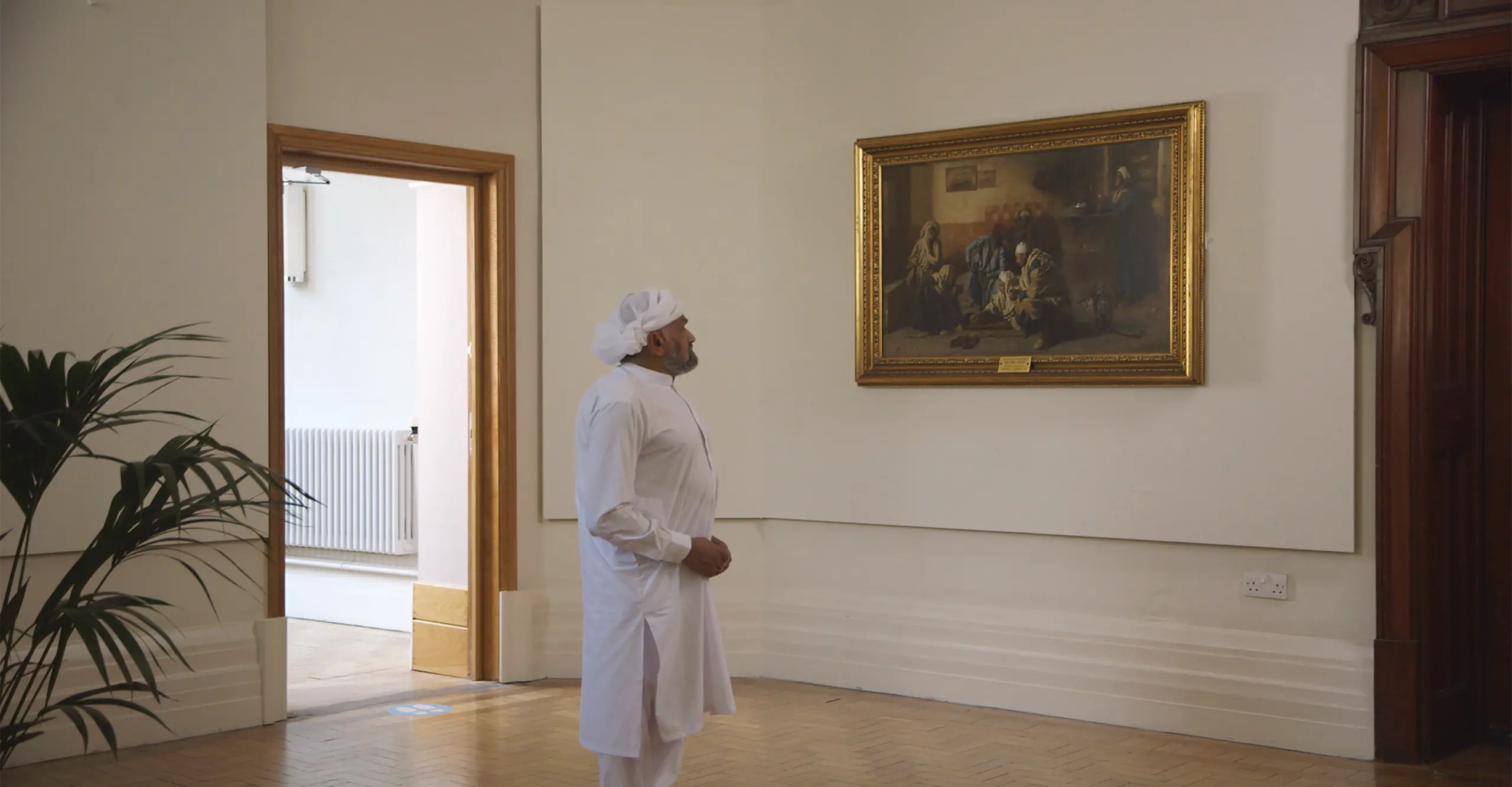 A man dressed in a white shalwar kameez and head covering stands in front of a painting in a gallery.