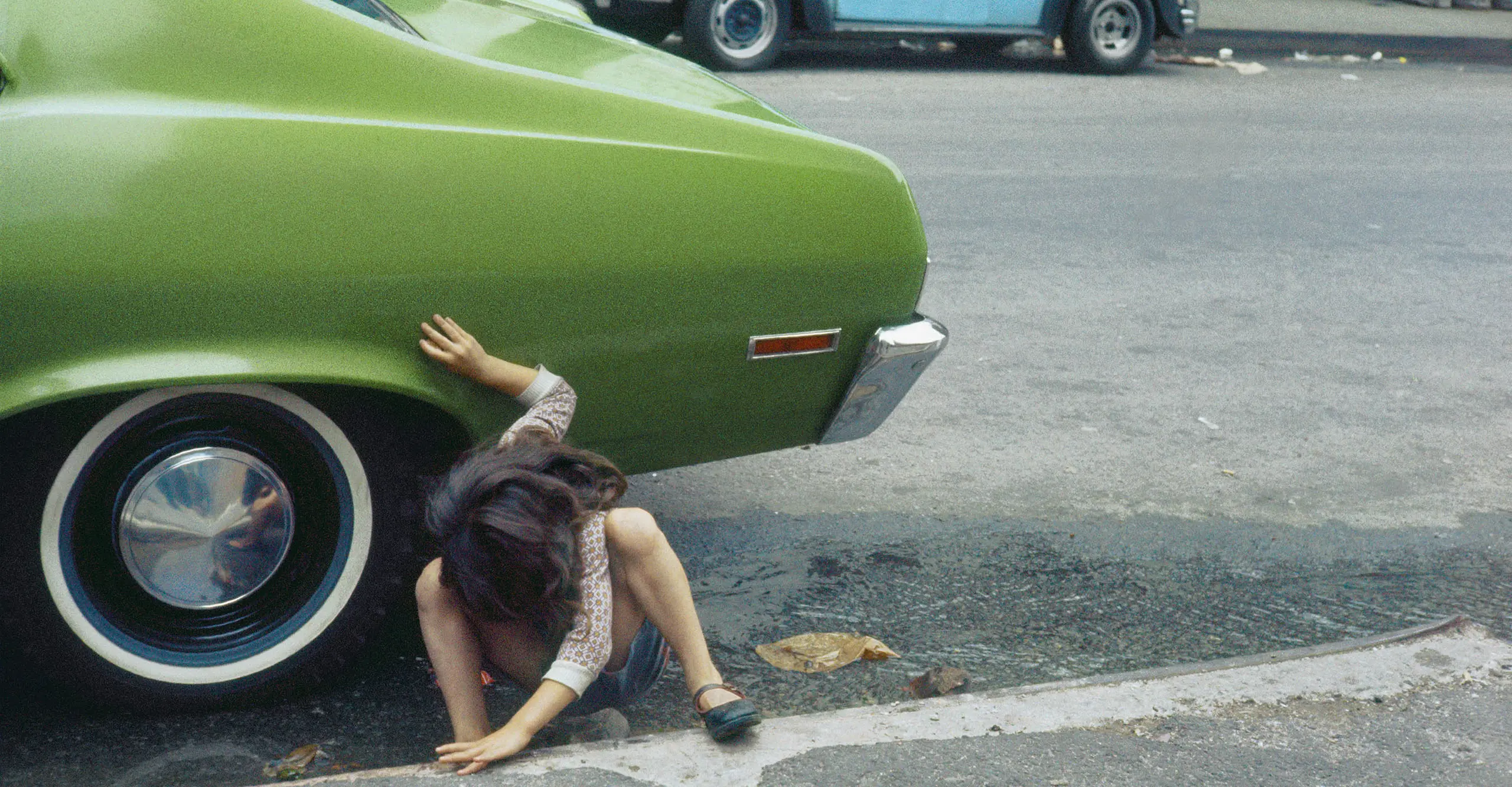 A photograph of the back end of a green car with a person crouched down near the wheel.