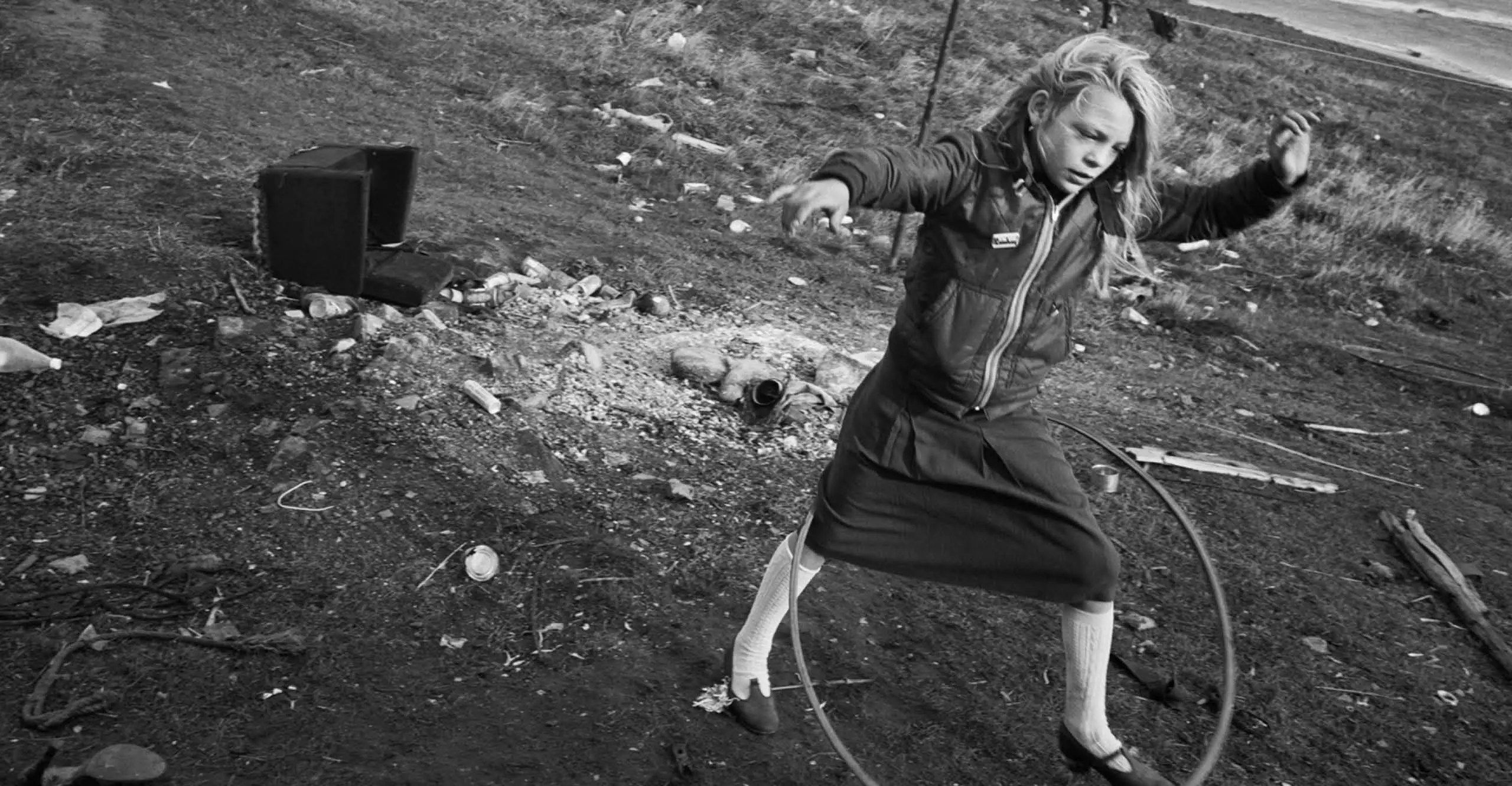 Black and white photograph of a girl playing with a hula-hoop, against a deindustrialized landscape