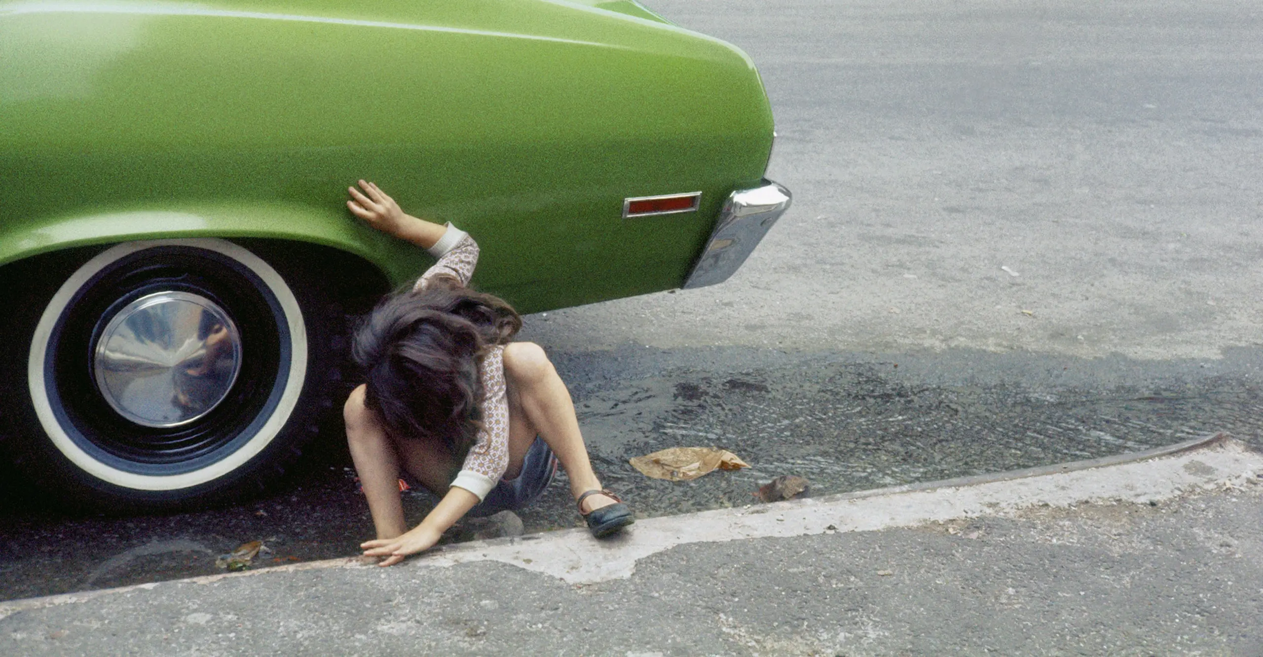 A child crouches on the pavement next to the back tyre of a green car