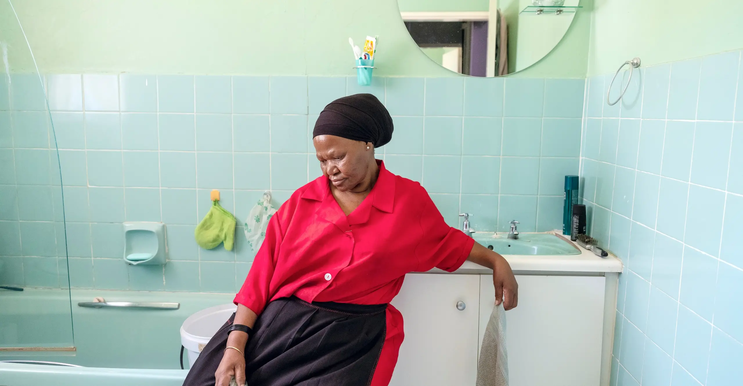 Colour photograph of a woman sat in a bathroom looking down. She holds a sponge in her hand.