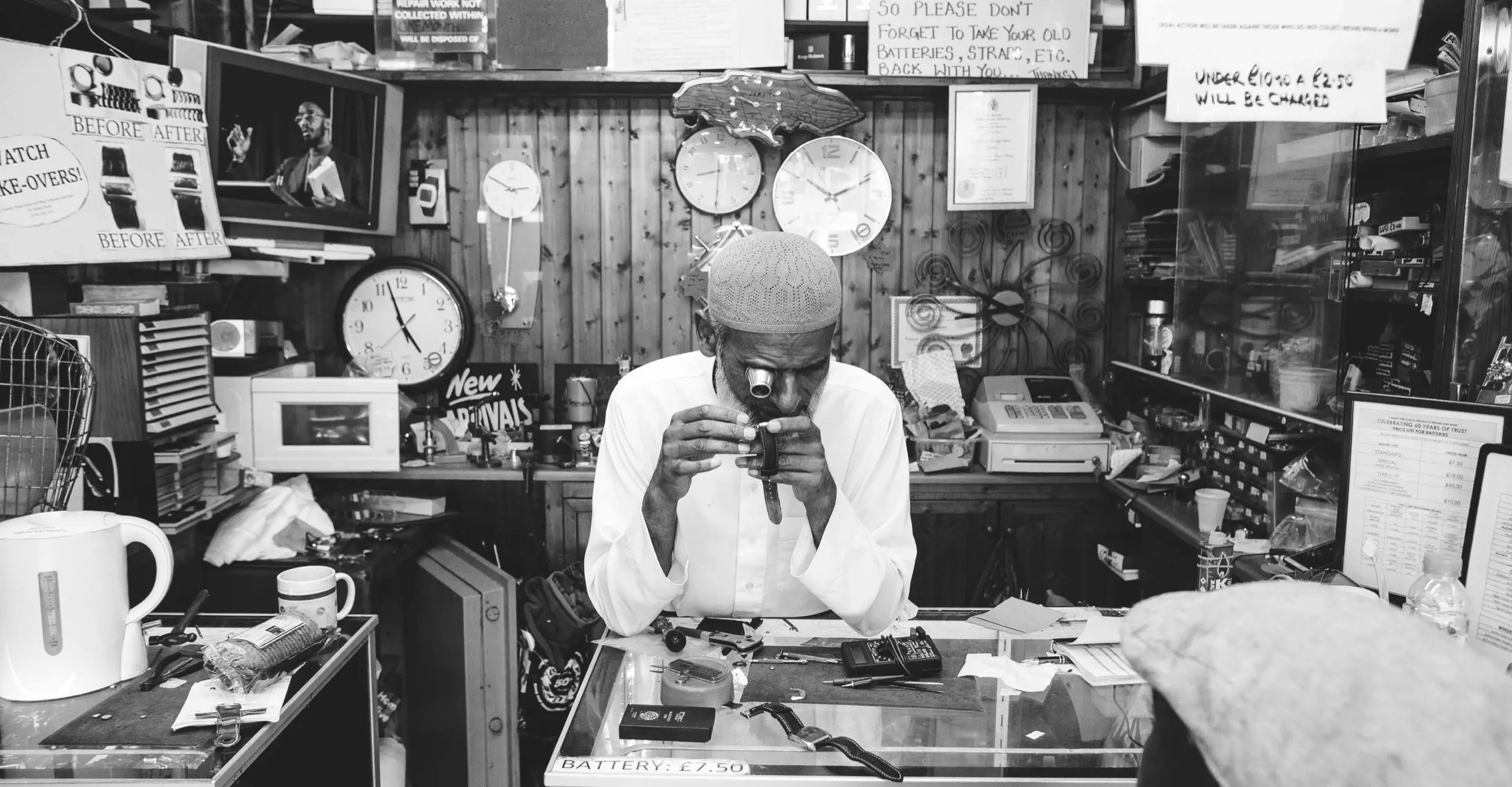 Black and white image featuring a man in a shop leaning on a vitrine surrounded by clocks