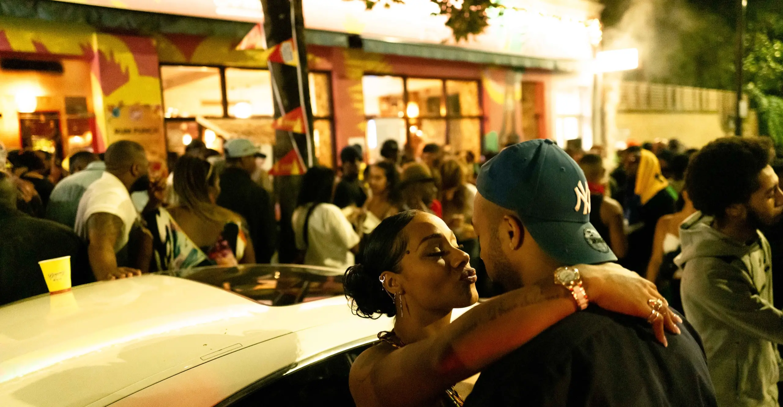 Candid photo of a couple kissing against a car with a crowd of people in the background