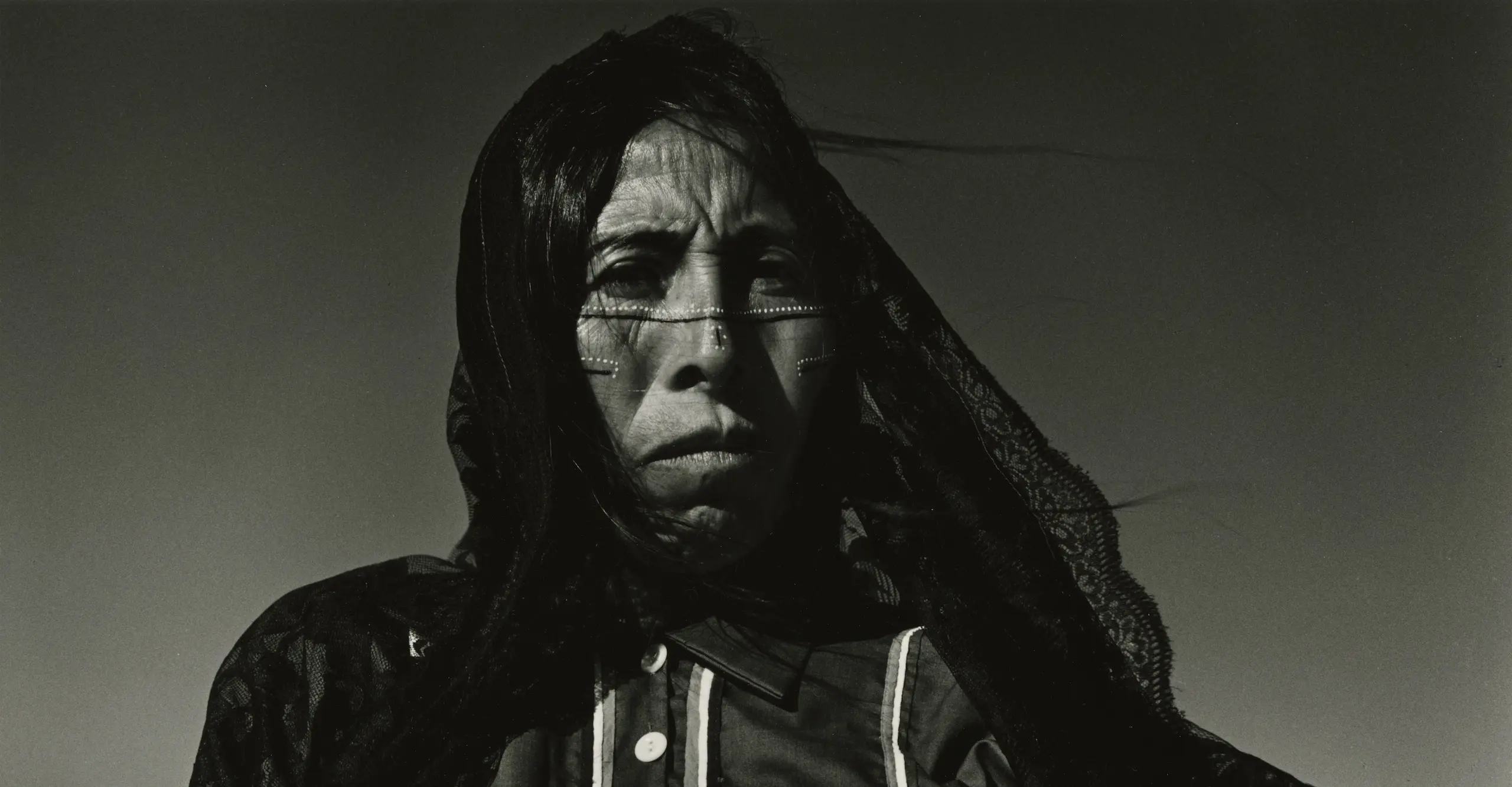 Black and white portrait photograph of a woman with indigenous paintings on her face and a black veil over her head.