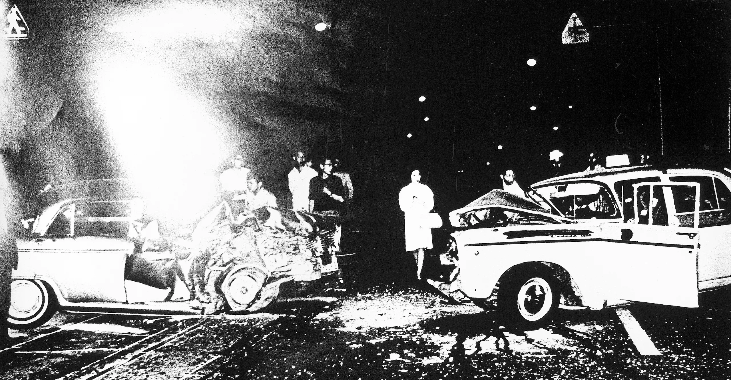 High contrast black and white photograph of two severely damaged cars and a small crowd