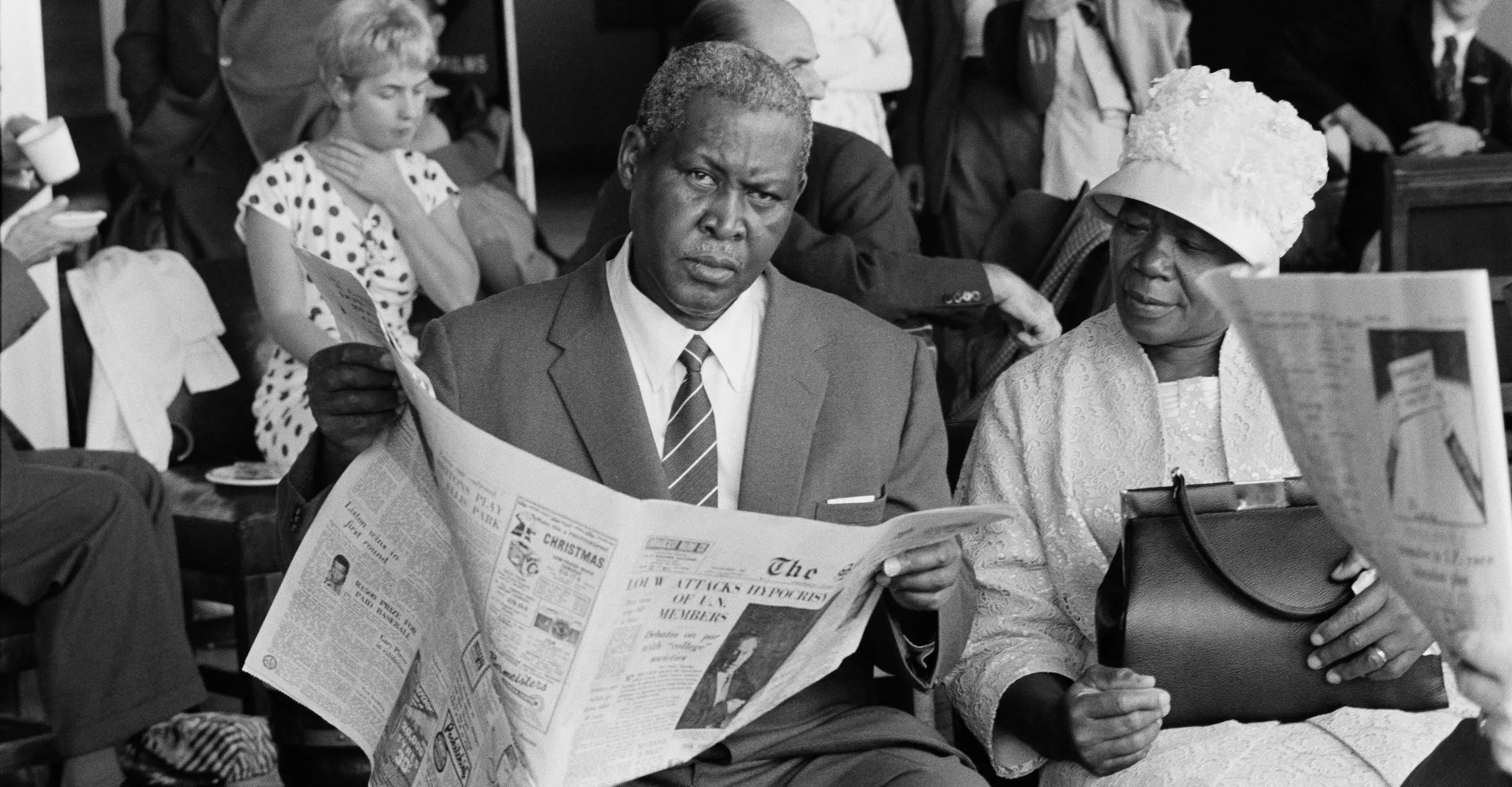 Black and white photograph of Albert Luthuli sat with a newspaper in his hands, as he looks direct-ly into the camera. His wife is sat beside him.
