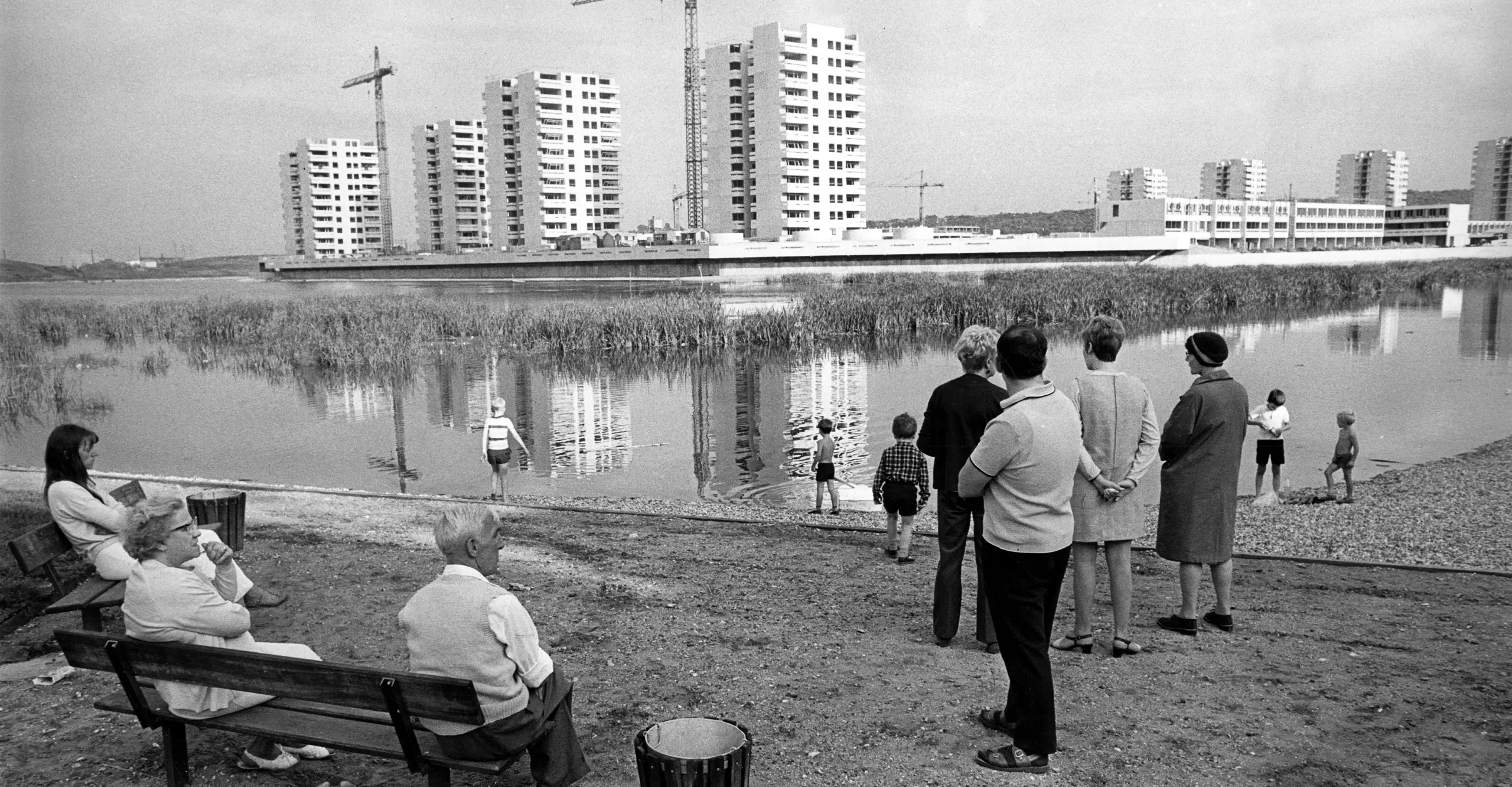 Black and white photograph of group of people on river bank with apartments being constructed in background